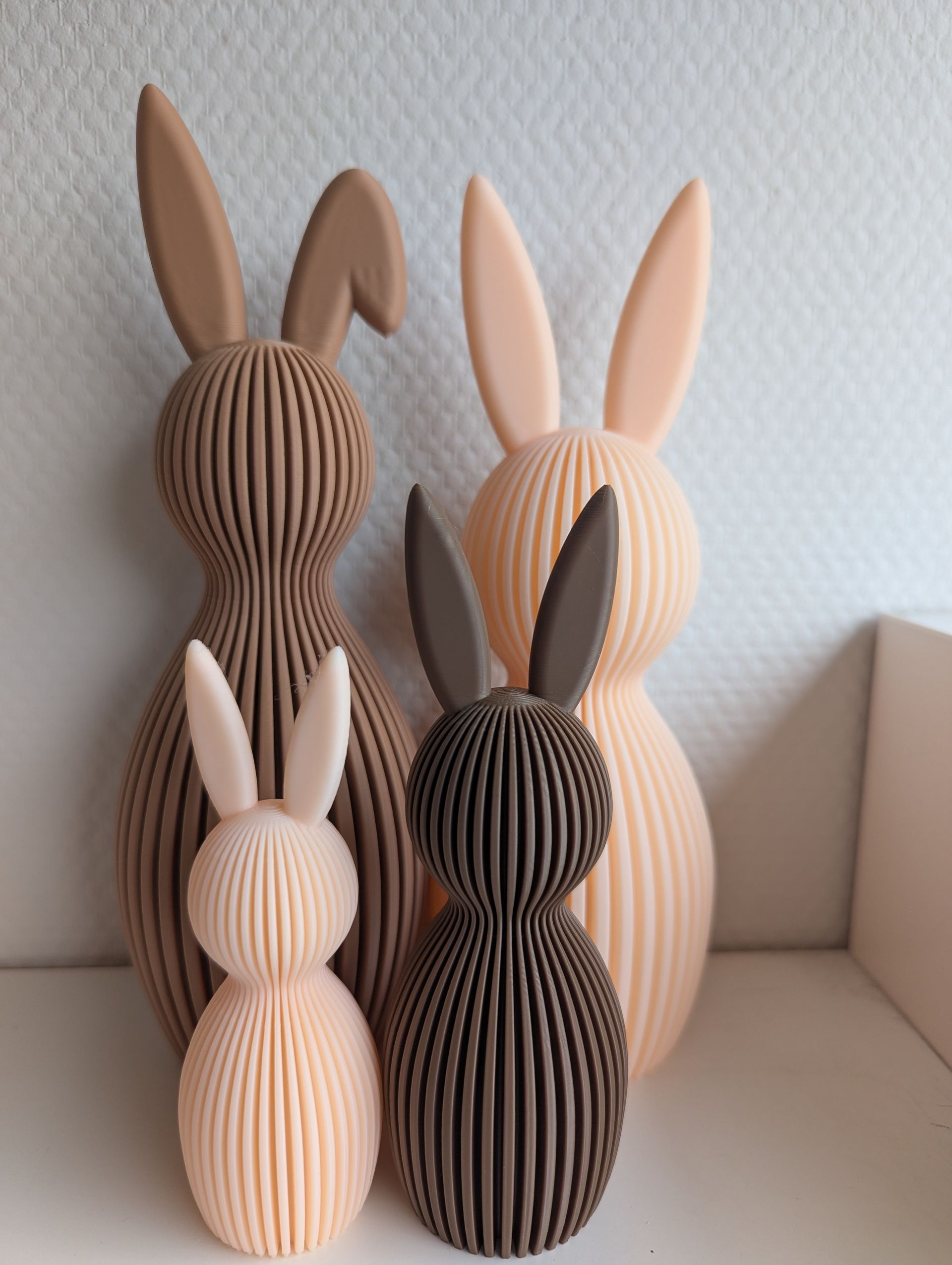 Rig-3d Vier geribbelde, konijnvormige decoratieve figuren in afwisselend licht perzik- en donkerbruine kleuren staan tegen een witte muur.