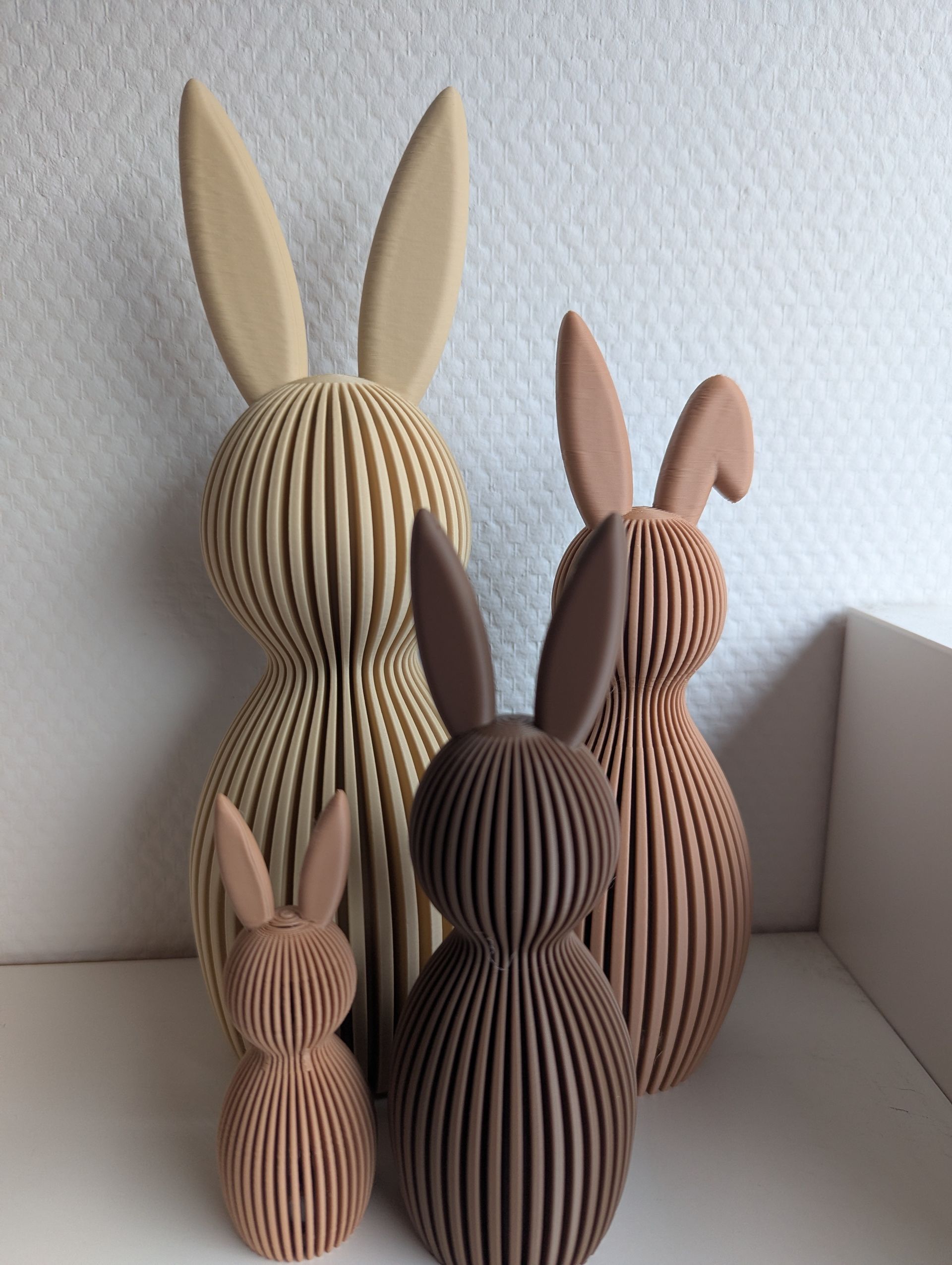 Rig-3d Vier geribbelde, konijnvormige decoraties in verschillende maten en bruintinten staan naast elkaar tegen een witte muur.