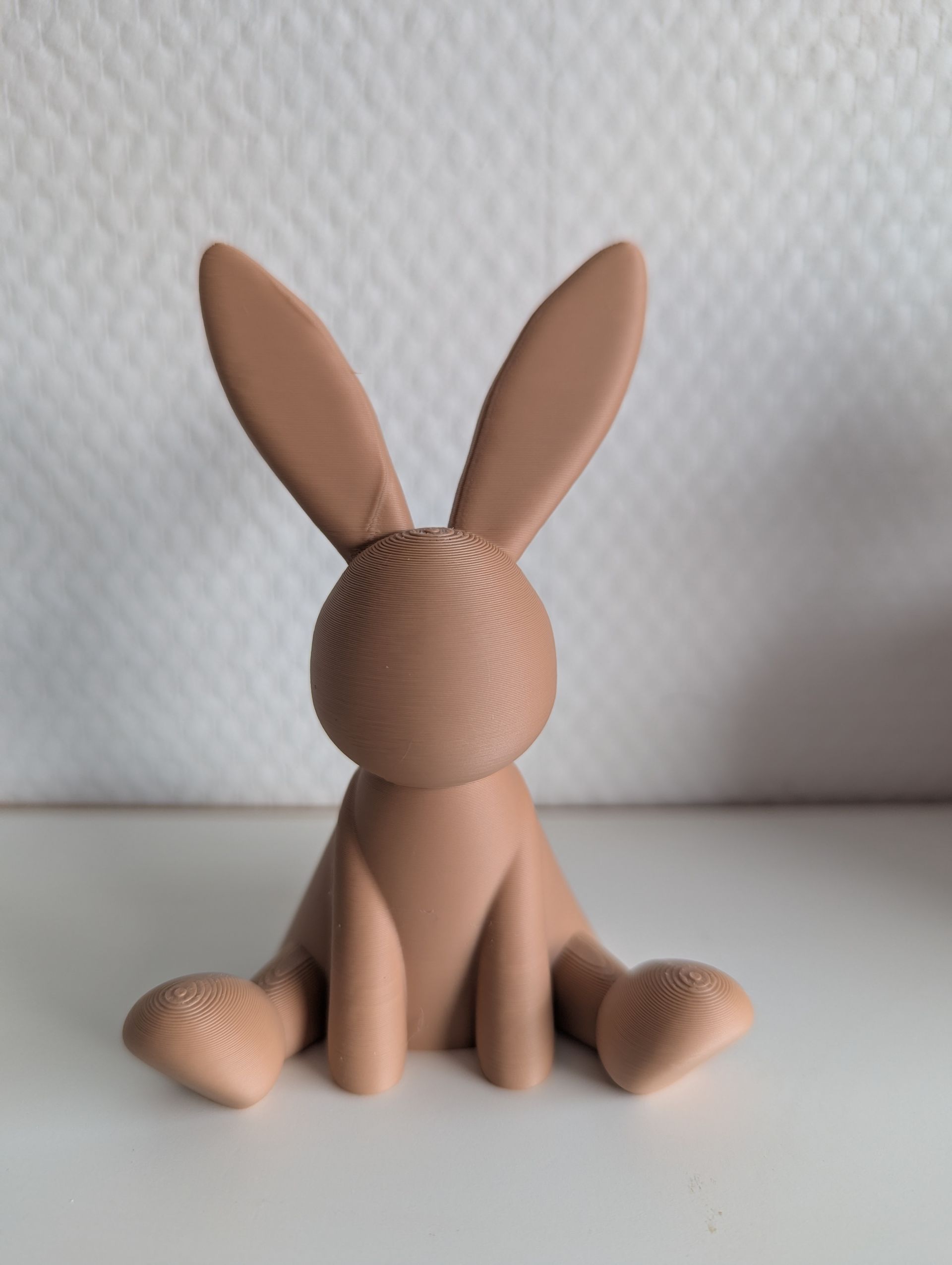 Rig-3d Een lichtbruin, 3D-geprint konijnenbeeldje dat op een witte ondergrond tegen een witte muur staat.