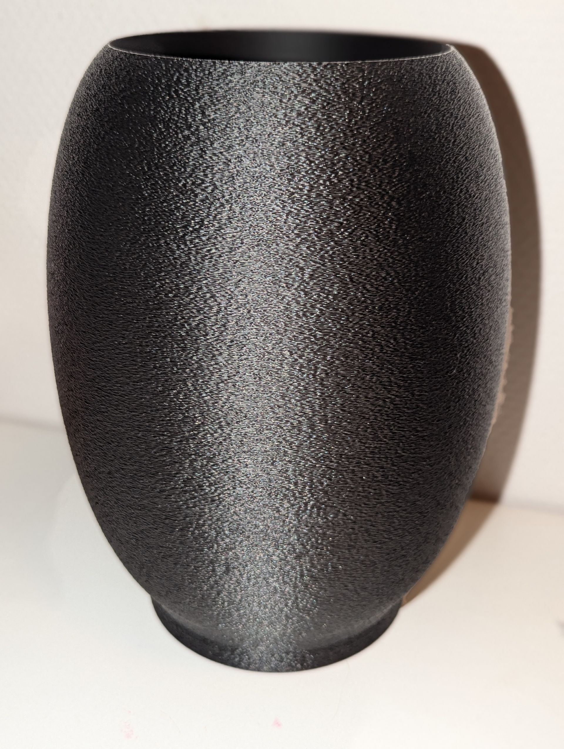 Rig-3d Een donkergrijs, glinsterend, vaasvormig 3D-geprint object met een gestructureerd oppervlak, dat op een effen witte ondergrond staat.