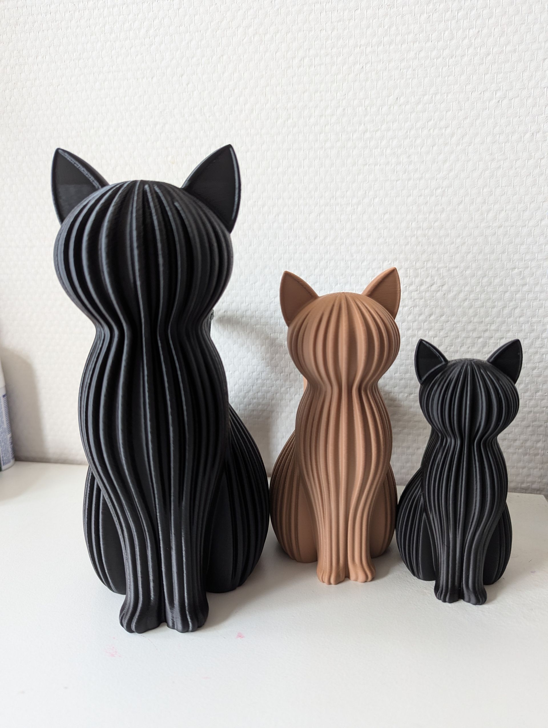 Rig-3d Drie kattenbeeldjes met een geribbelde textuur, gerangschikt van groot naar klein, op een witte achtergrond tegen een muur.