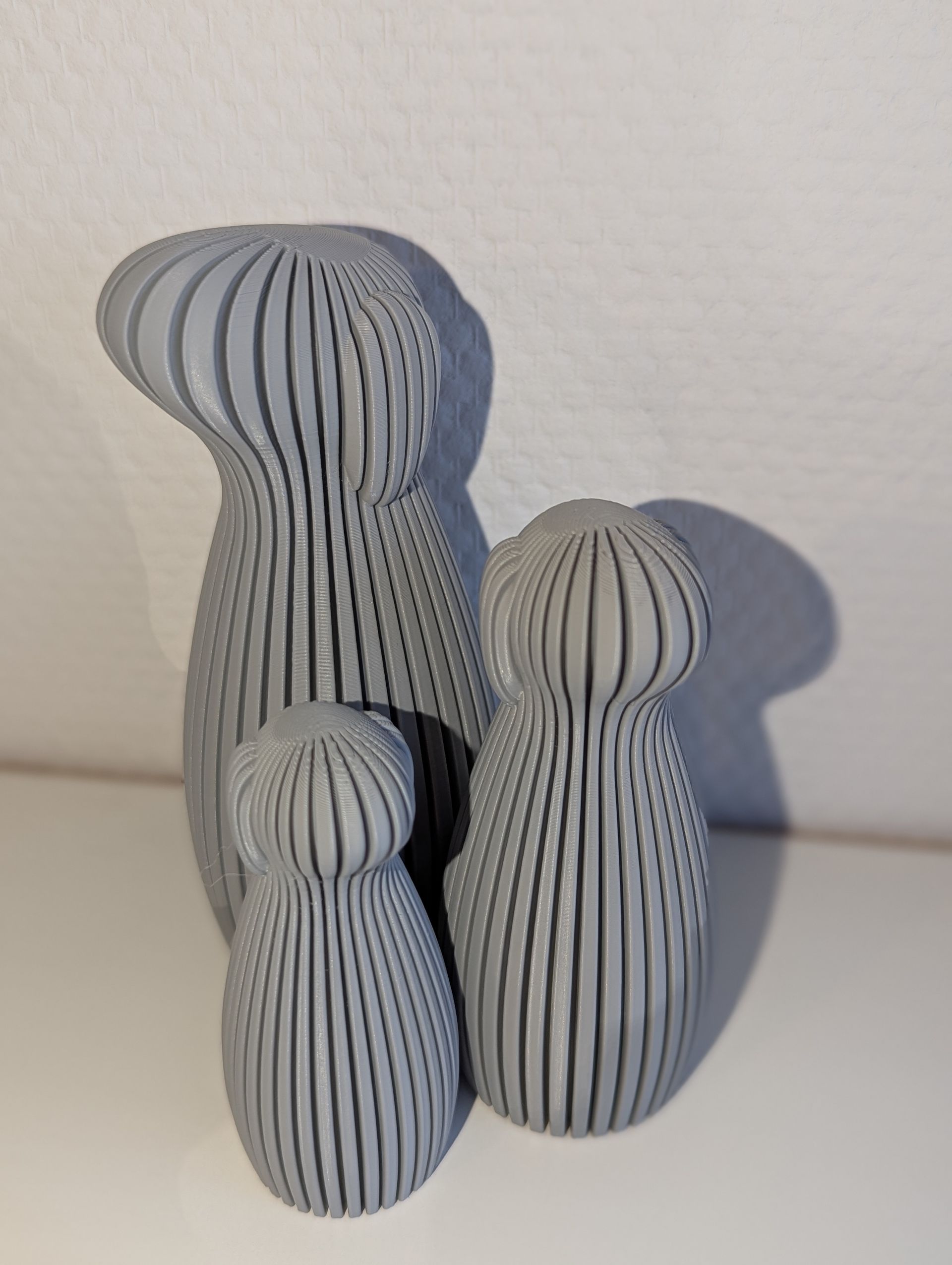 Rig-3d Drie grijze, verticaal geribbelde, afgeronde, 3D-geprinte abstracte sculpturen van verschillende afmetingen op een witte ondergrond.