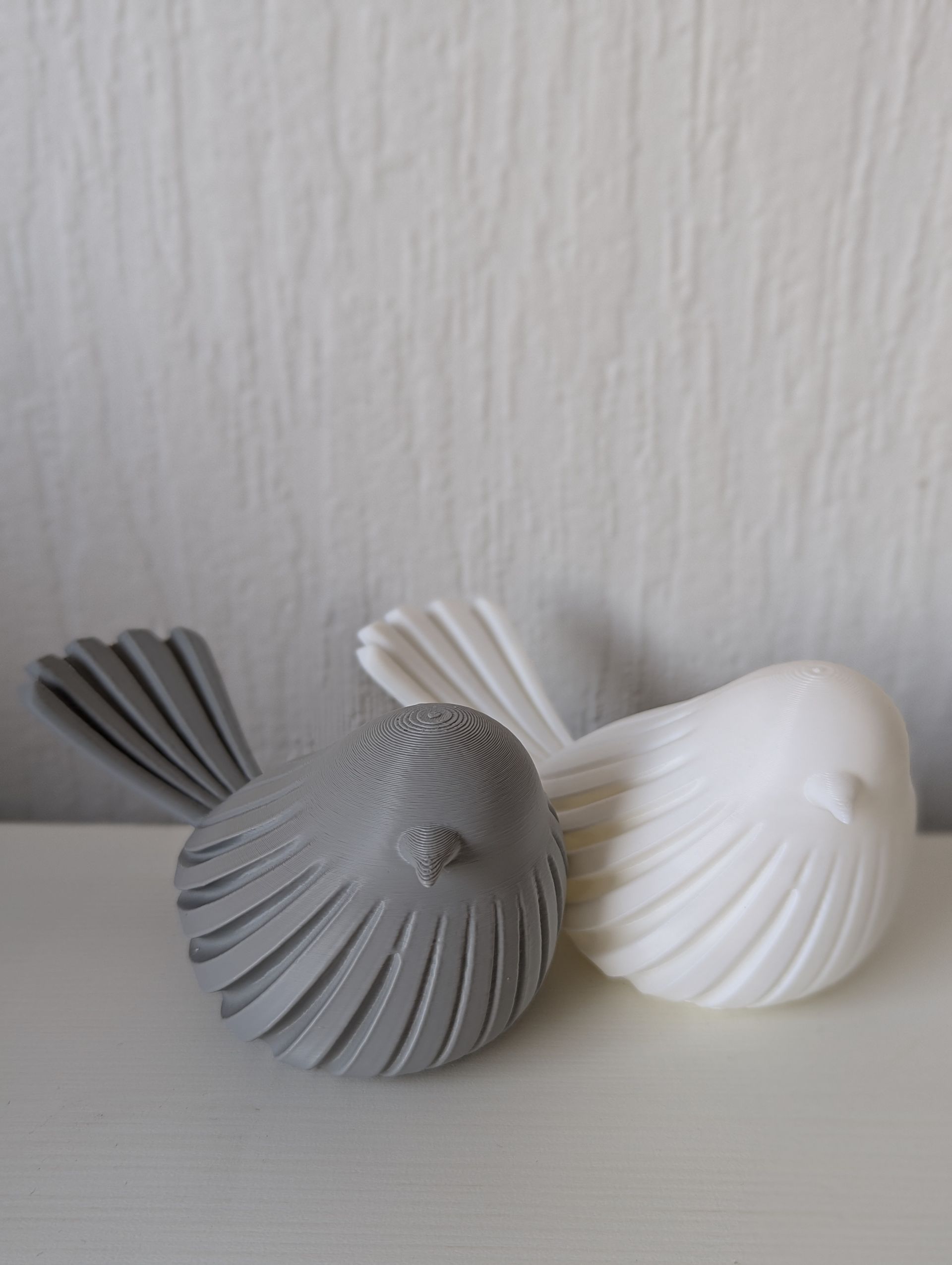 Rig-3d Twee kleine, gestileerde vogelbeeldjes – een grijs met een gedecoreerde kop, een wit – staan naast elkaar op een witte ondergrond.