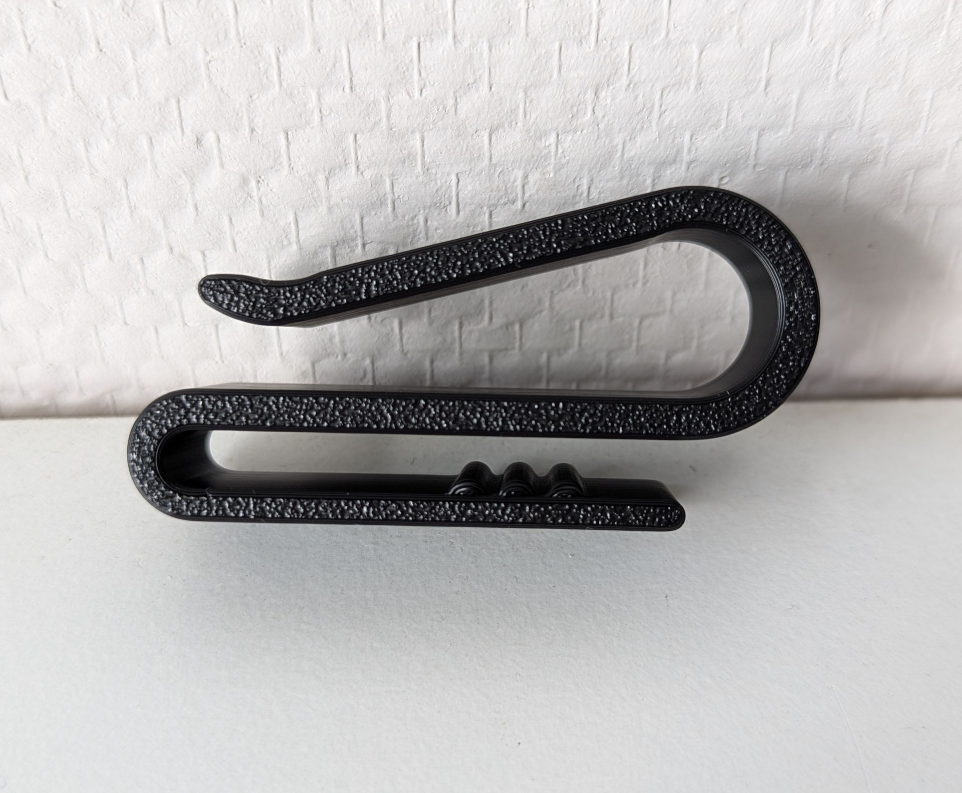 Rig-3d Een zwarte, S-vormige plastic riemclip met een gestructureerd oppervlak, rustend op een witte ondergrond tegen een witte muur.