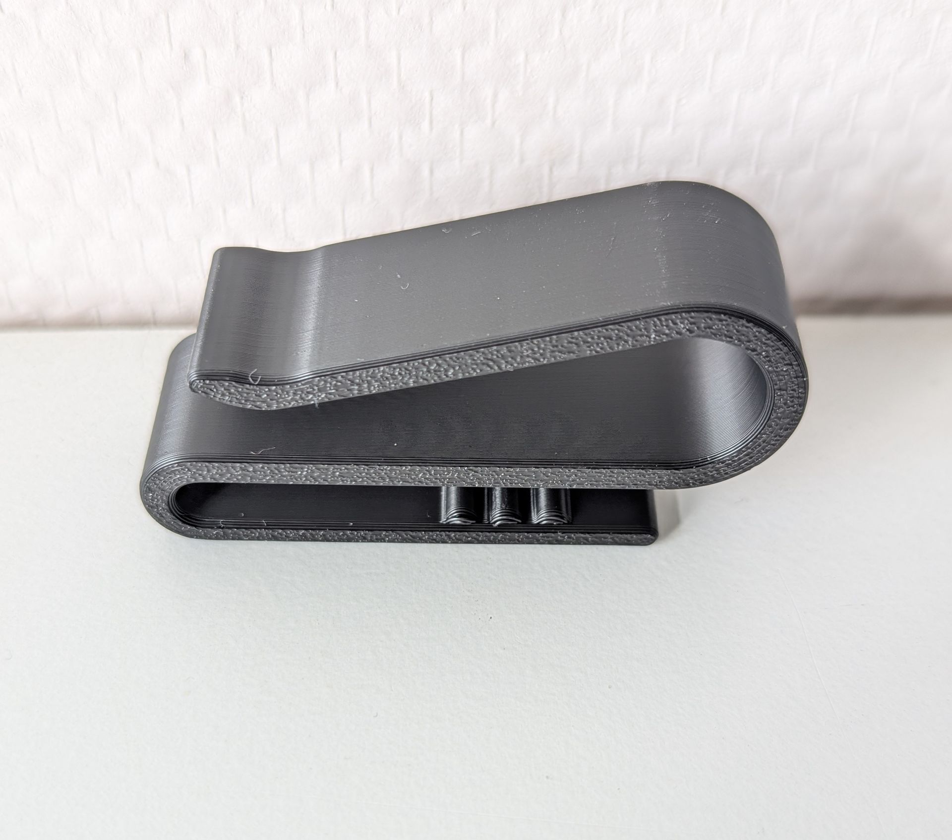 Rig-3d Een zwarte, S-vormige plastic riemclip met een gestructureerd oppervlak, rustend op een witte ondergrond tegen een witte muur.