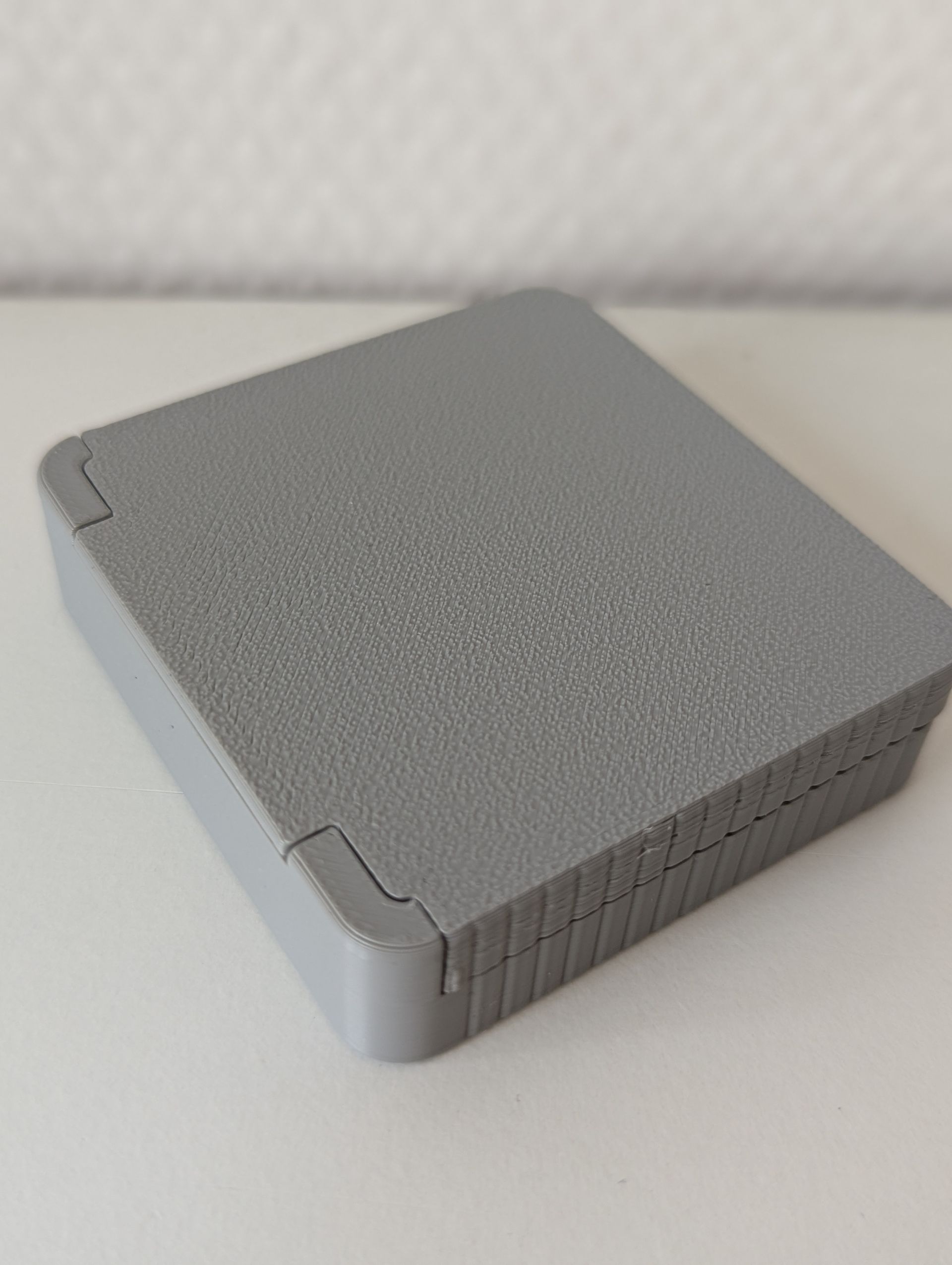 Rig-3d waterdrop Een grijze, vierkante 3D-geprinte doos met een gestructureerd bovenoppervlak, die op een vlakke witte ondergrond staat.