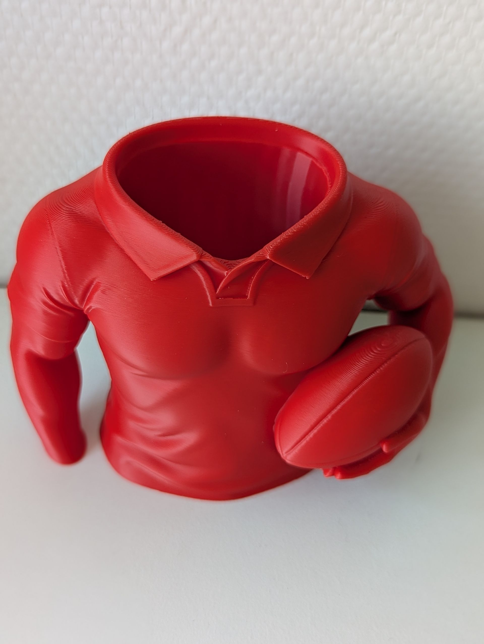 Rig-3d Een rode, 3D-geprinte bureau-organizer in de vorm van een gespierd bovenlichaam met een poloshirt aan en een rugbybal in zijn handen.