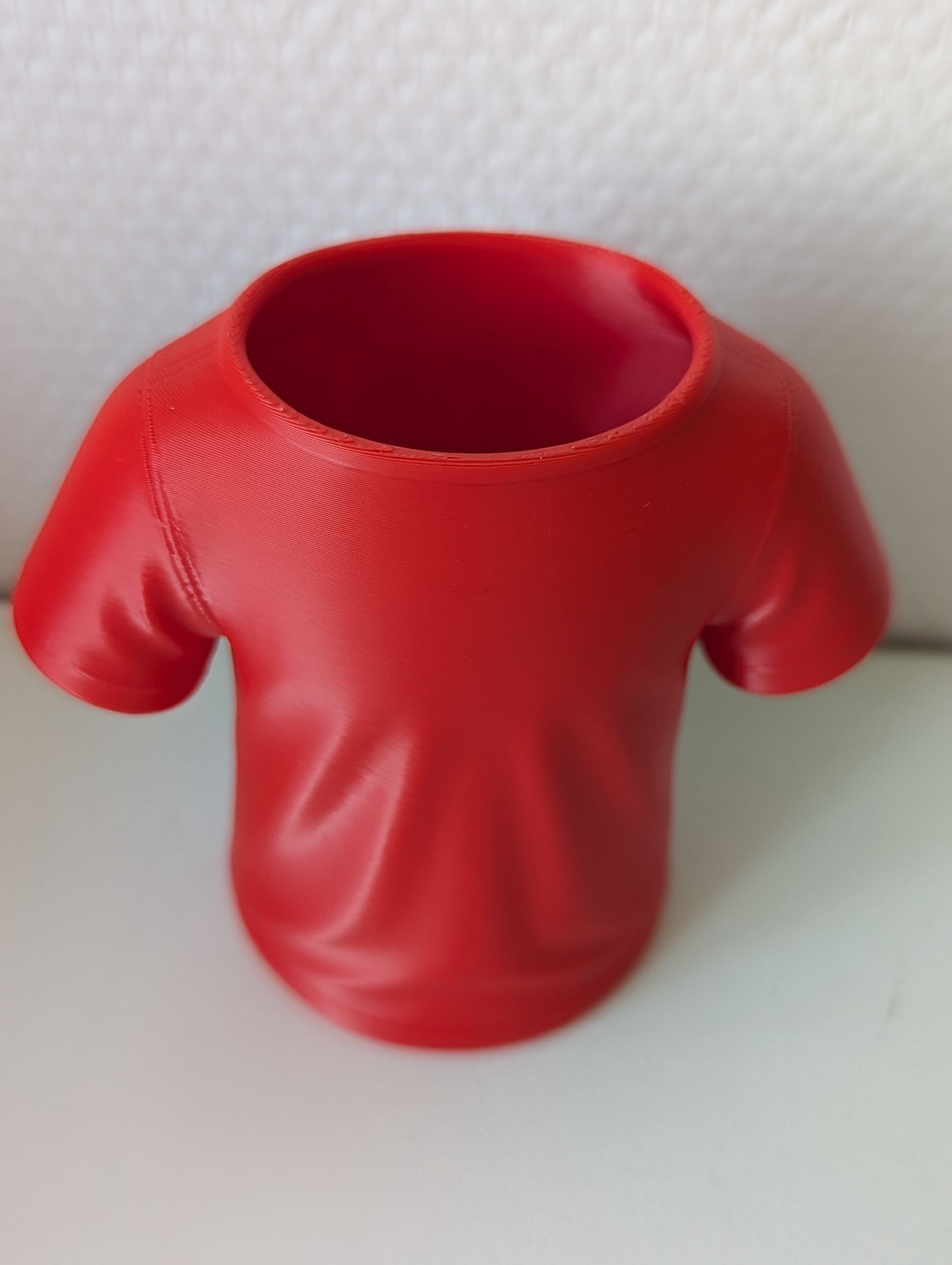 Rig-3d Een 3D-geprinte, rode T-shirtvormige houder of pennenhouder die op een vlakke, witte ondergrond staat.