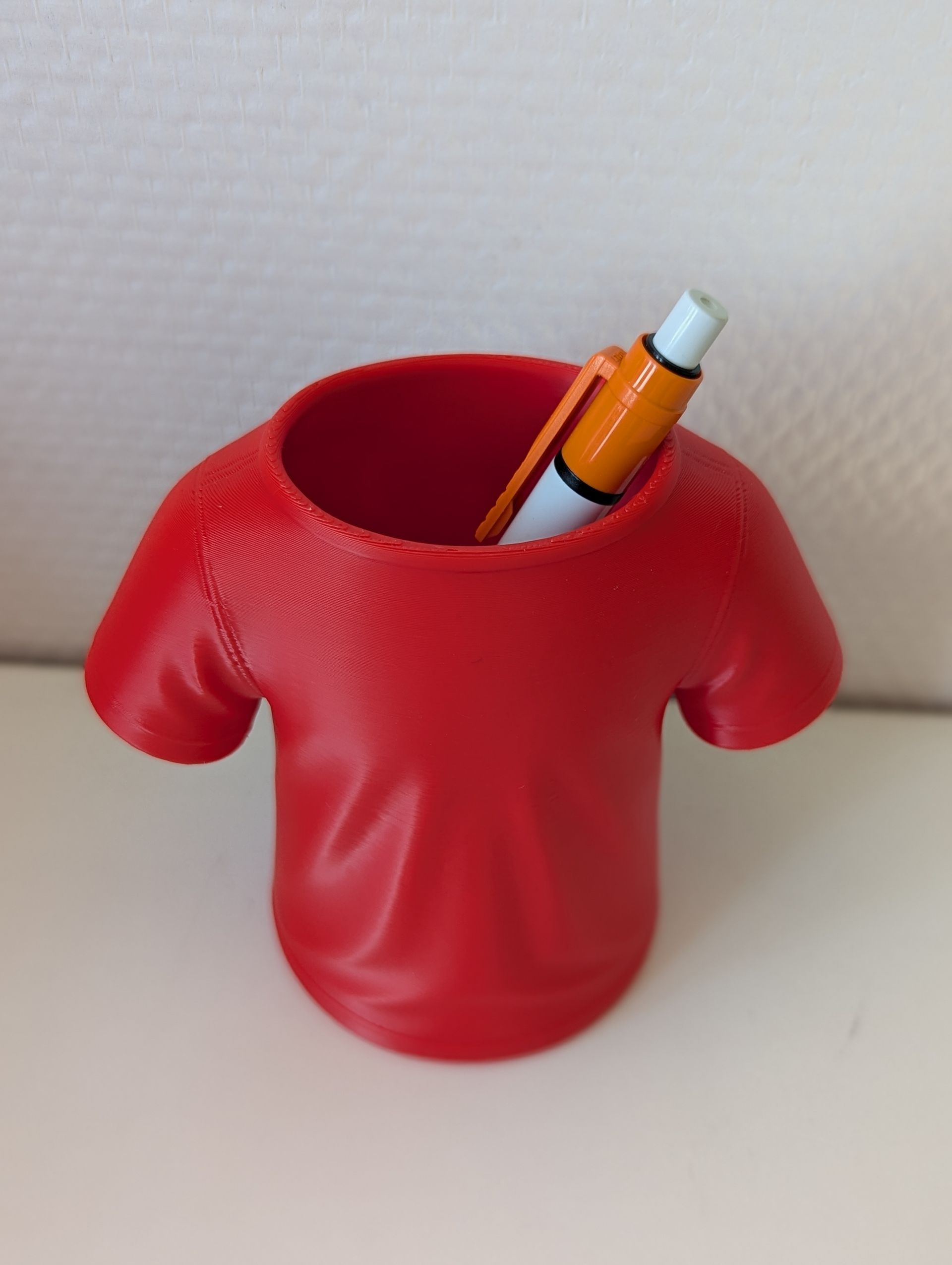 Rig-3d Een 3D-geprinte, rode T-shirtvormige houder of pennenhouder die op een vlakke, witte ondergrond staat.