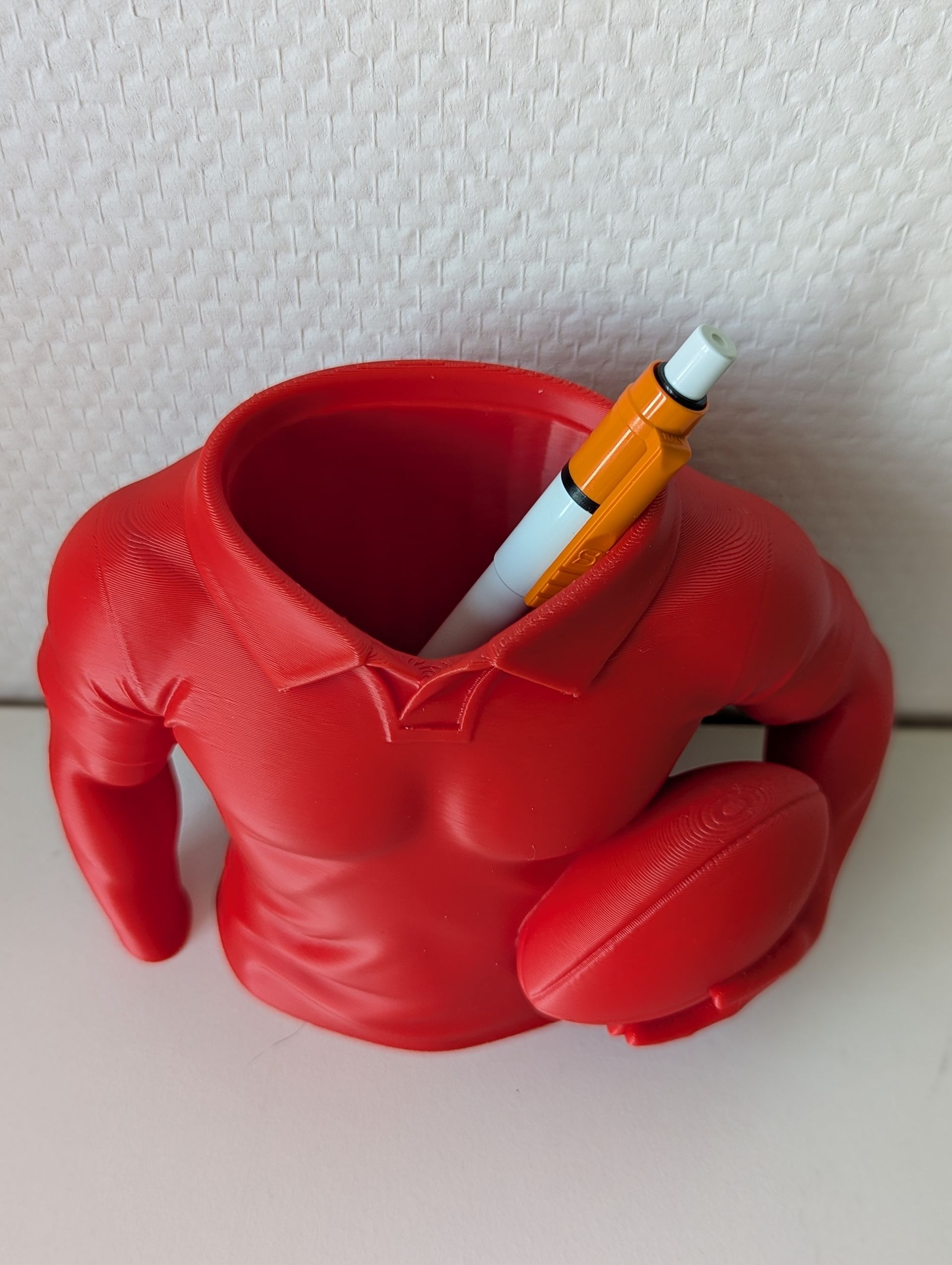 Rig-3d Een rode, 3D-geprinte bureau-organizer in de vorm van een gespierd bovenlichaam met een poloshirt aan en een rugbybal in zijn handen.