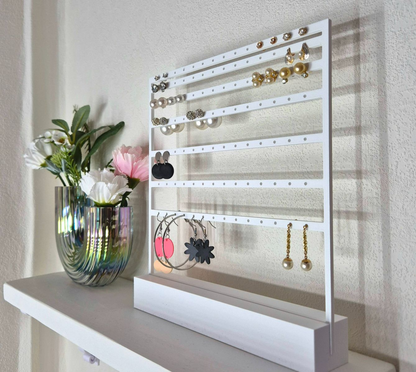 Rig-3d Een wit oorbellenhoudertje staat op een witte plank naast een vaas met bloemen tegen een lichte muur.