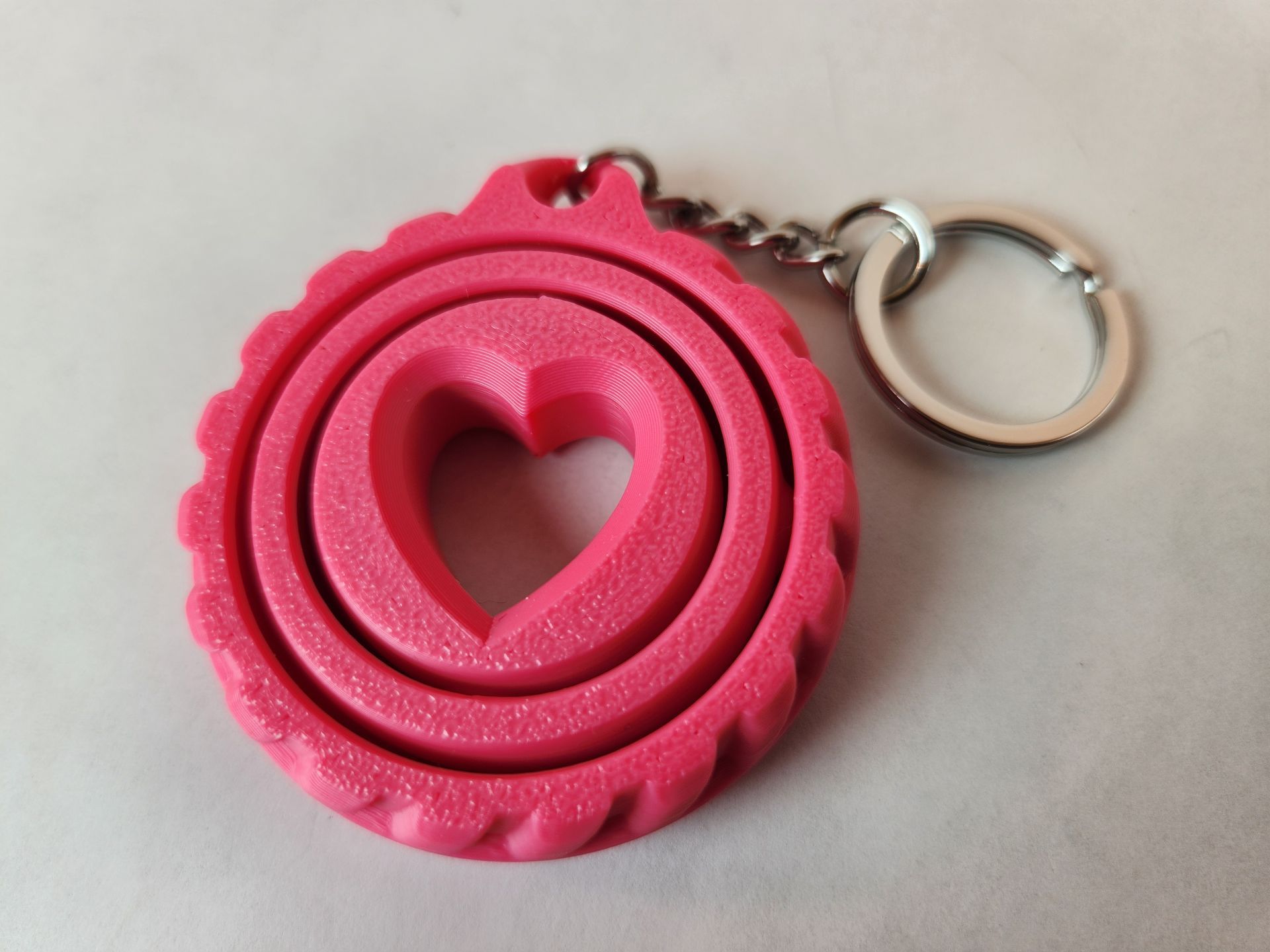 Rig-3d Een felroze, 3D-geprinte ronde hartvormige sleutelhanger met tandwielachtige randen, bevestigd aan een metalen splitring.