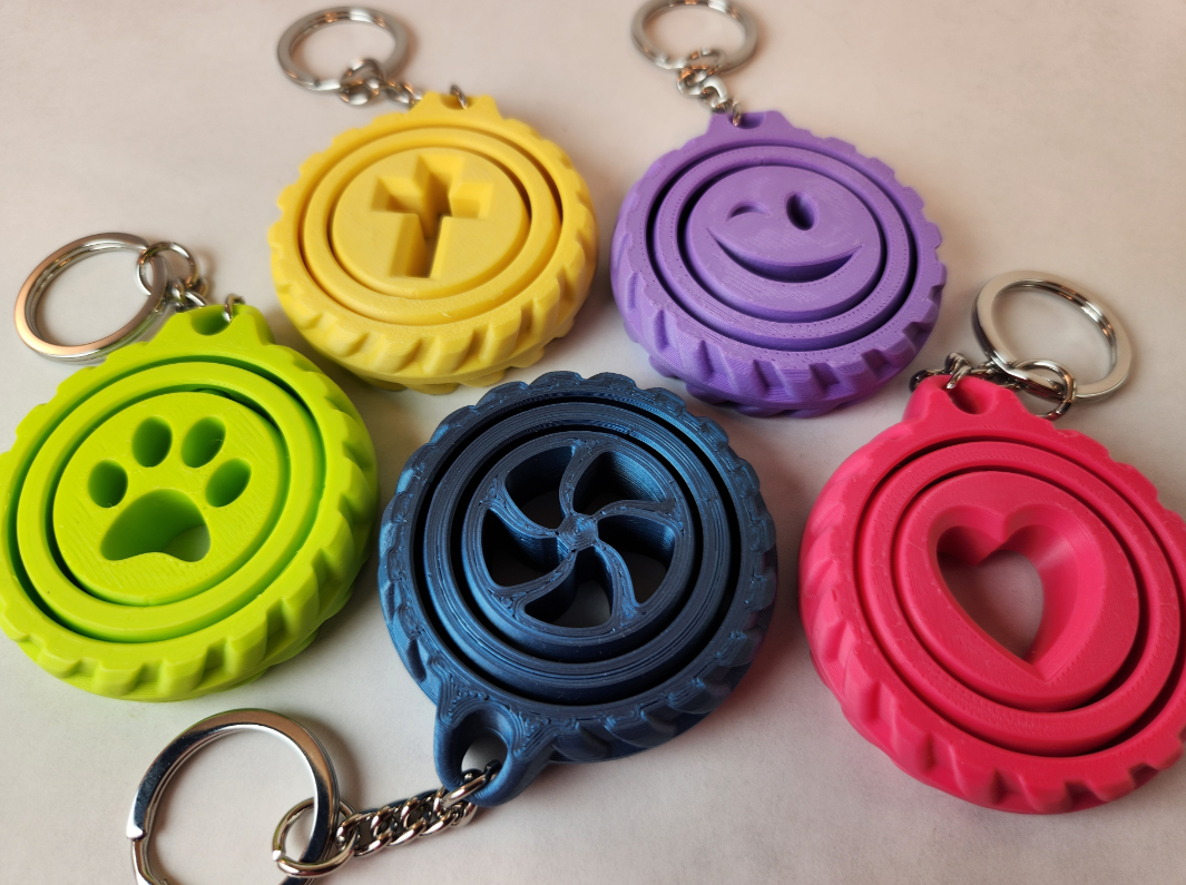 Rig-3d Vijf kleurrijke, 3D-geprinte ronde sleutelhangers met verschillende vormen in het midden: een pootafdruk, een kruis, een smiley, een spiraal en een hartje.