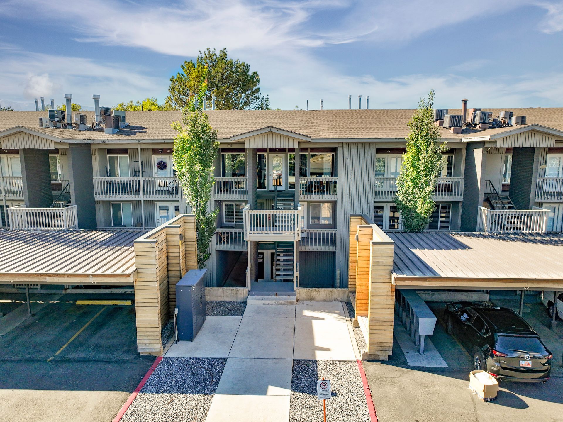 Apartment complex exterior with covered parking, balconies, and walkway. Gray building, blue sky.