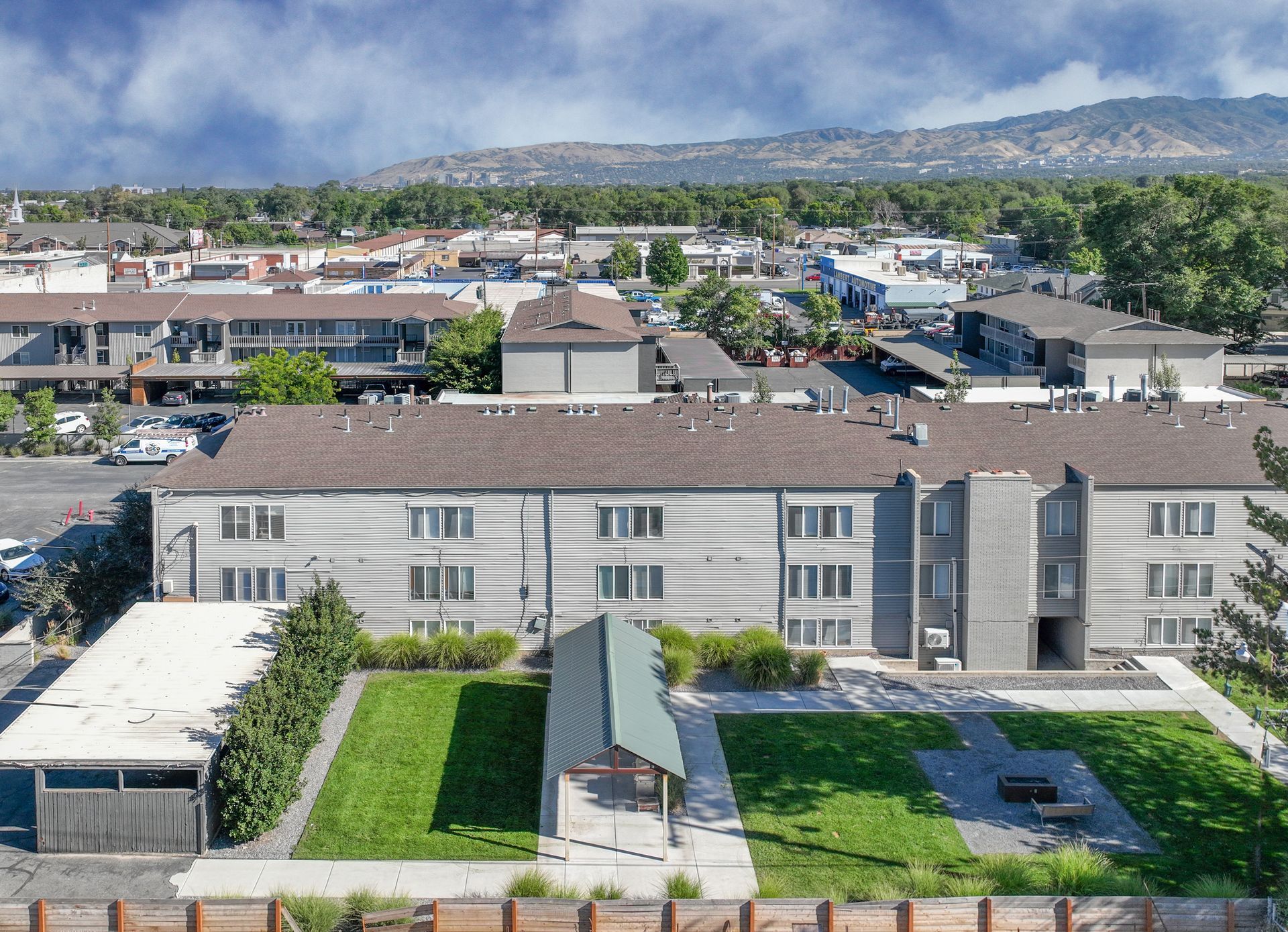 Aerial view of a gray apartment building with a green lawn, gazebo, and surrounding cityscape against a mountain backdrop.