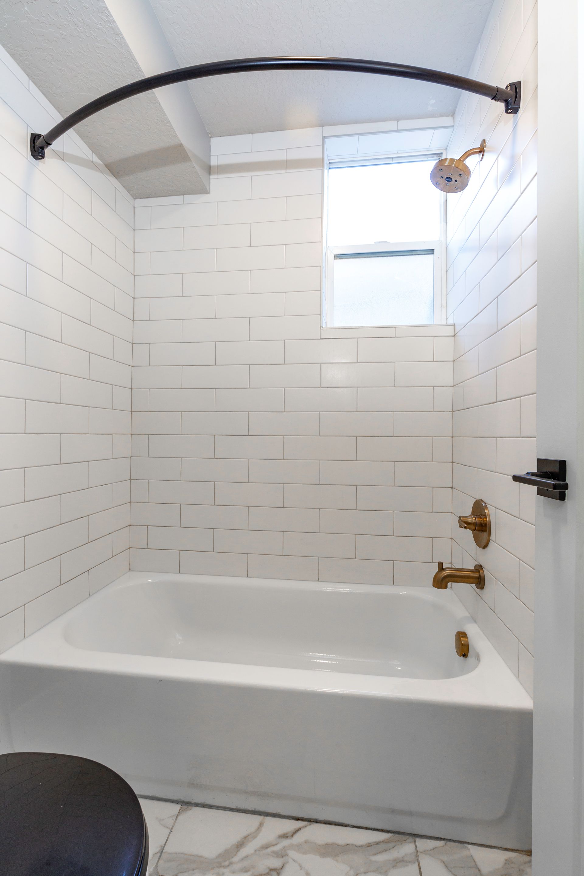 White-tiled bathroom with bathtub, gold fixtures, and a curved shower rod. A small window is in the wall.