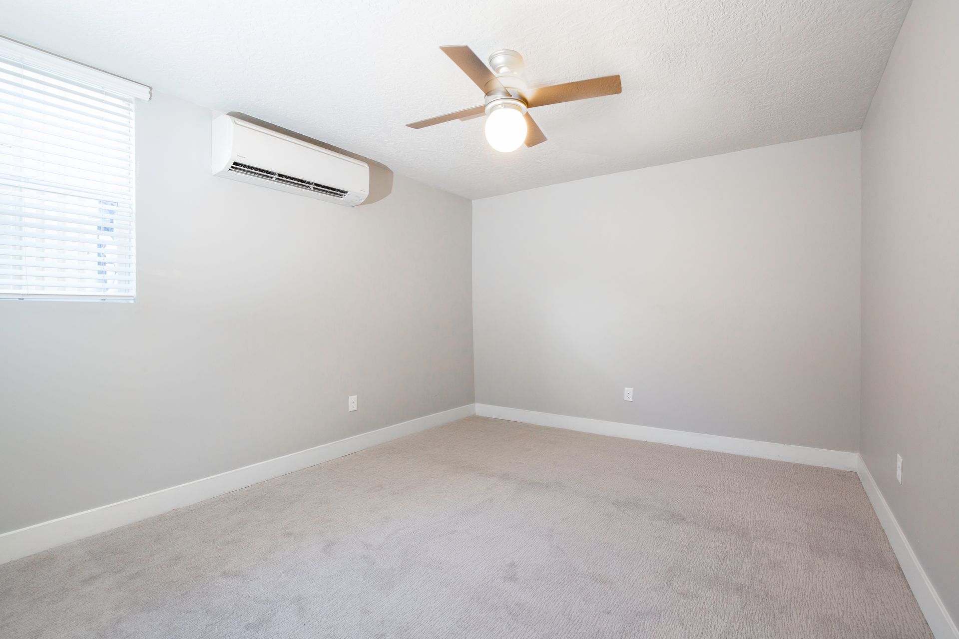 Empty room with gray walls and carpet; a window, air conditioner, and ceiling fan are visible.