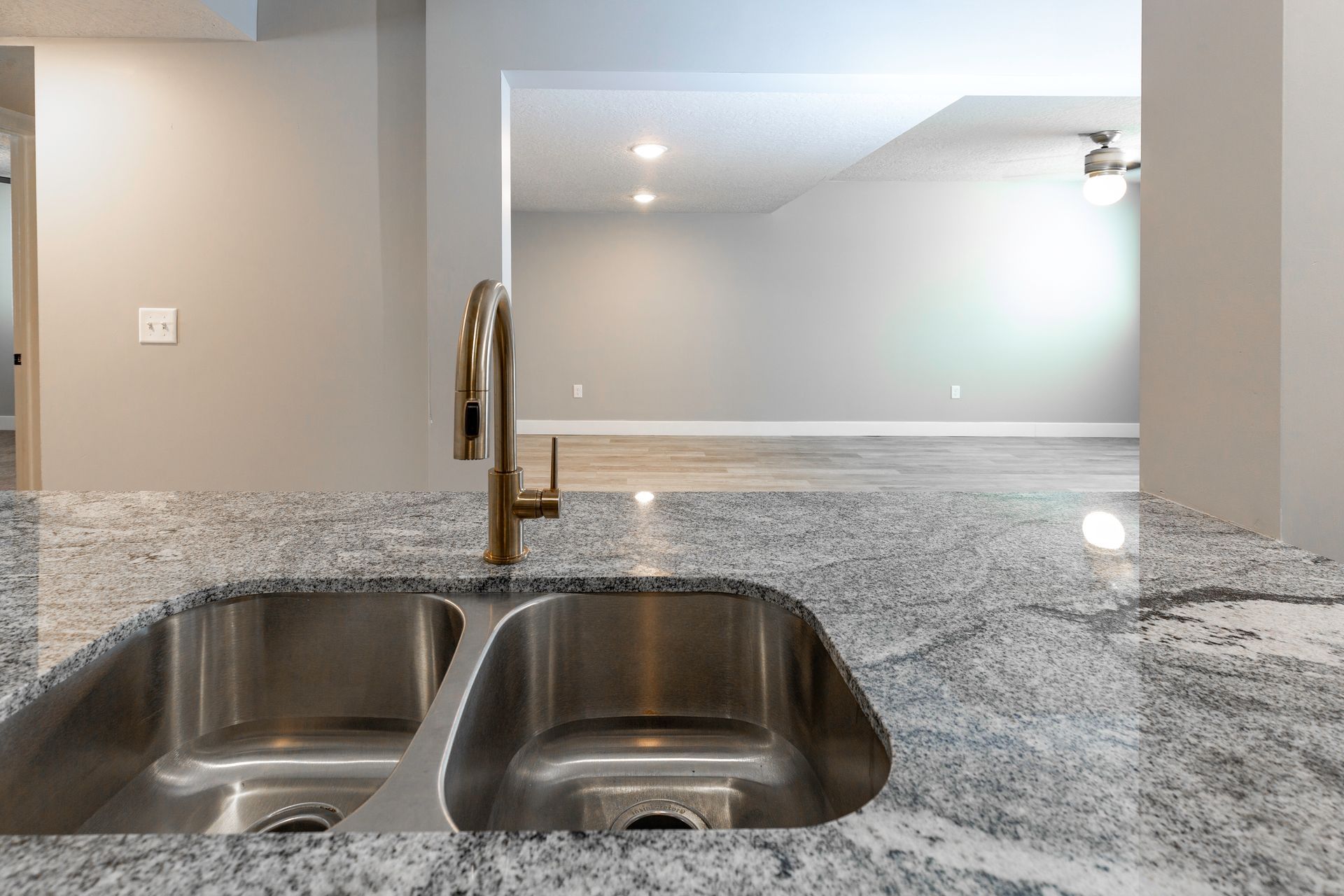 Stainless steel kitchen sink with gold faucet, granite countertop, leading to a living area.