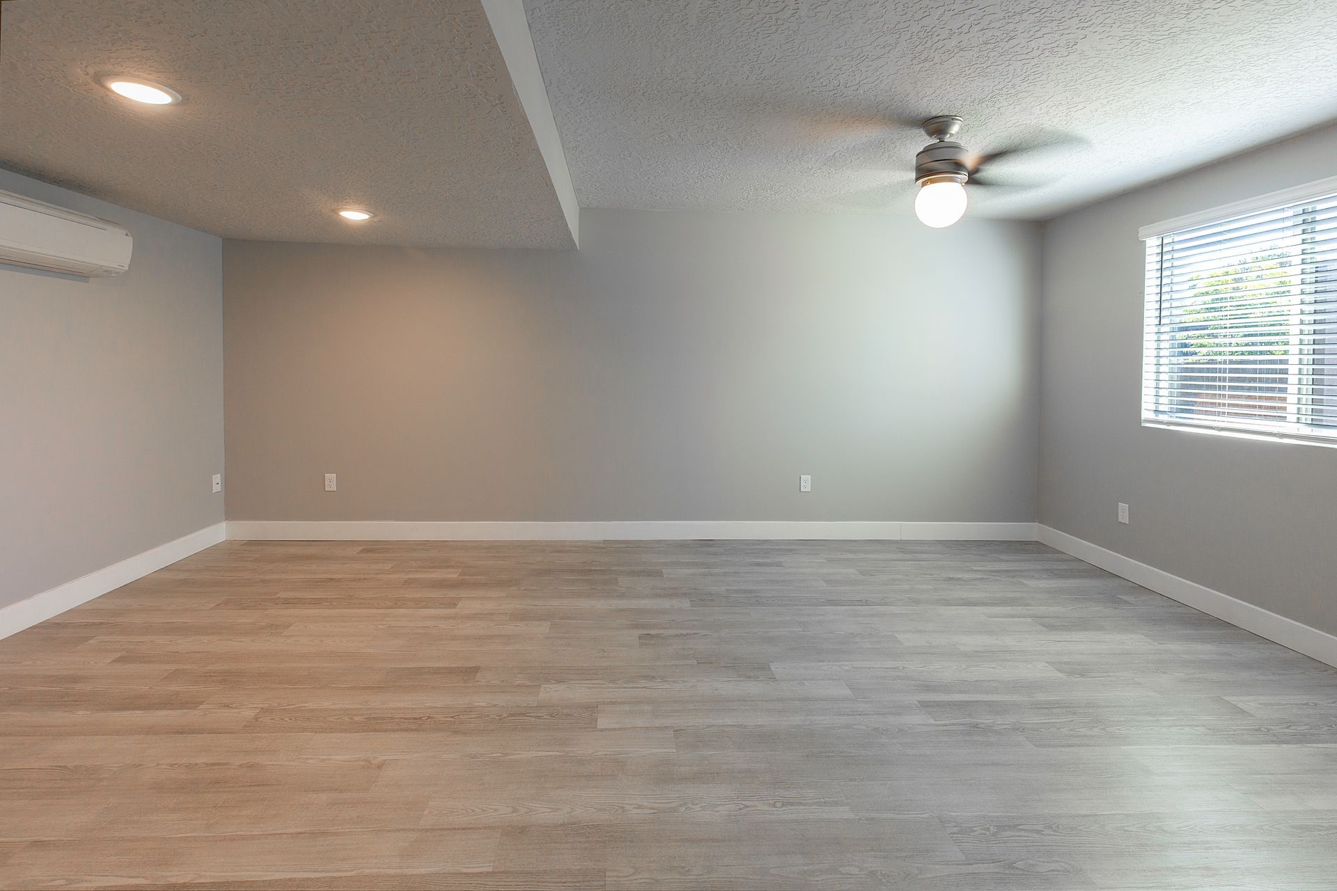 Empty room with gray walls, wooden floor, ceiling fan, and window with blinds.