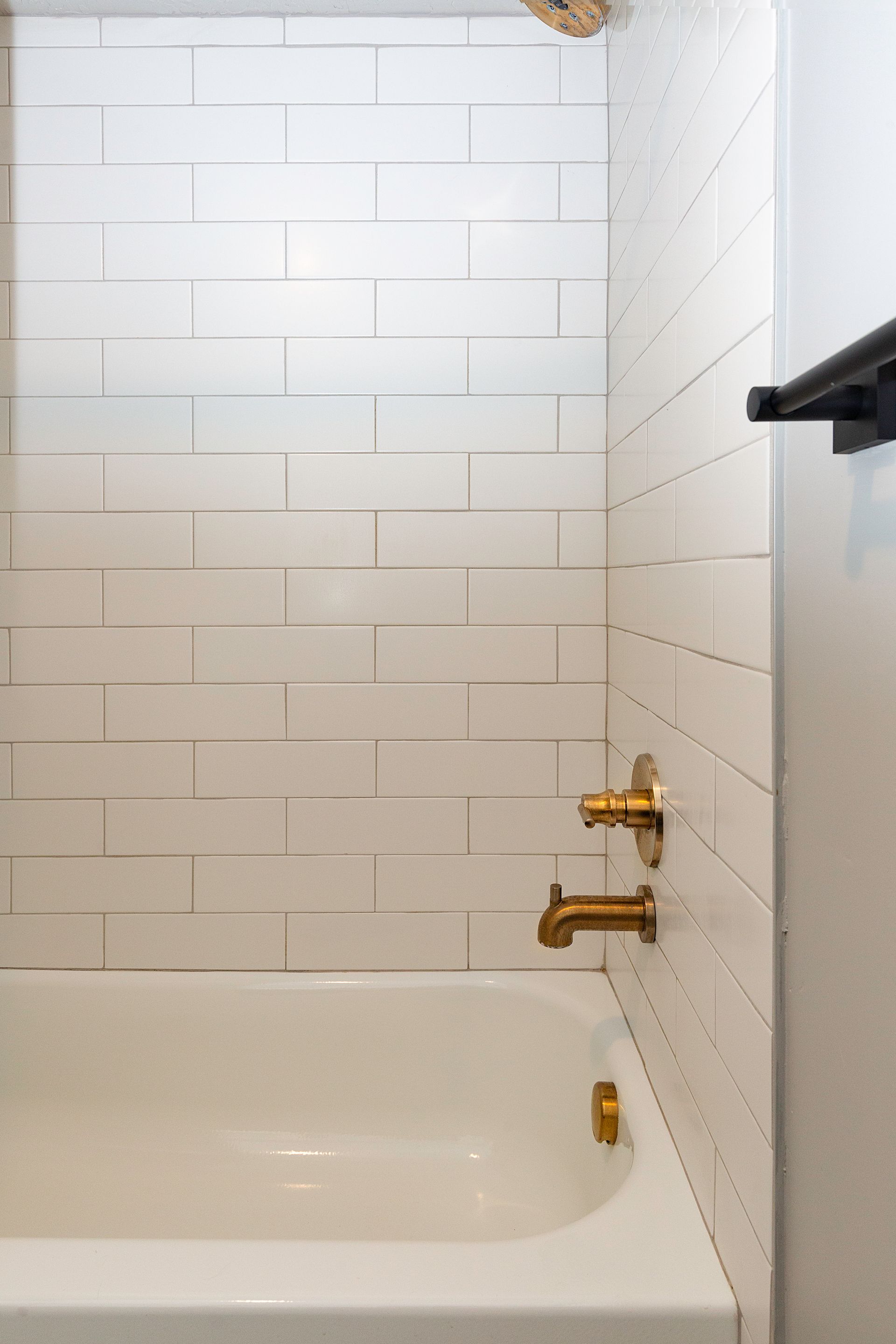 White-tiled shower with a gold faucet and a white bathtub. Black towel rack on the wall.
