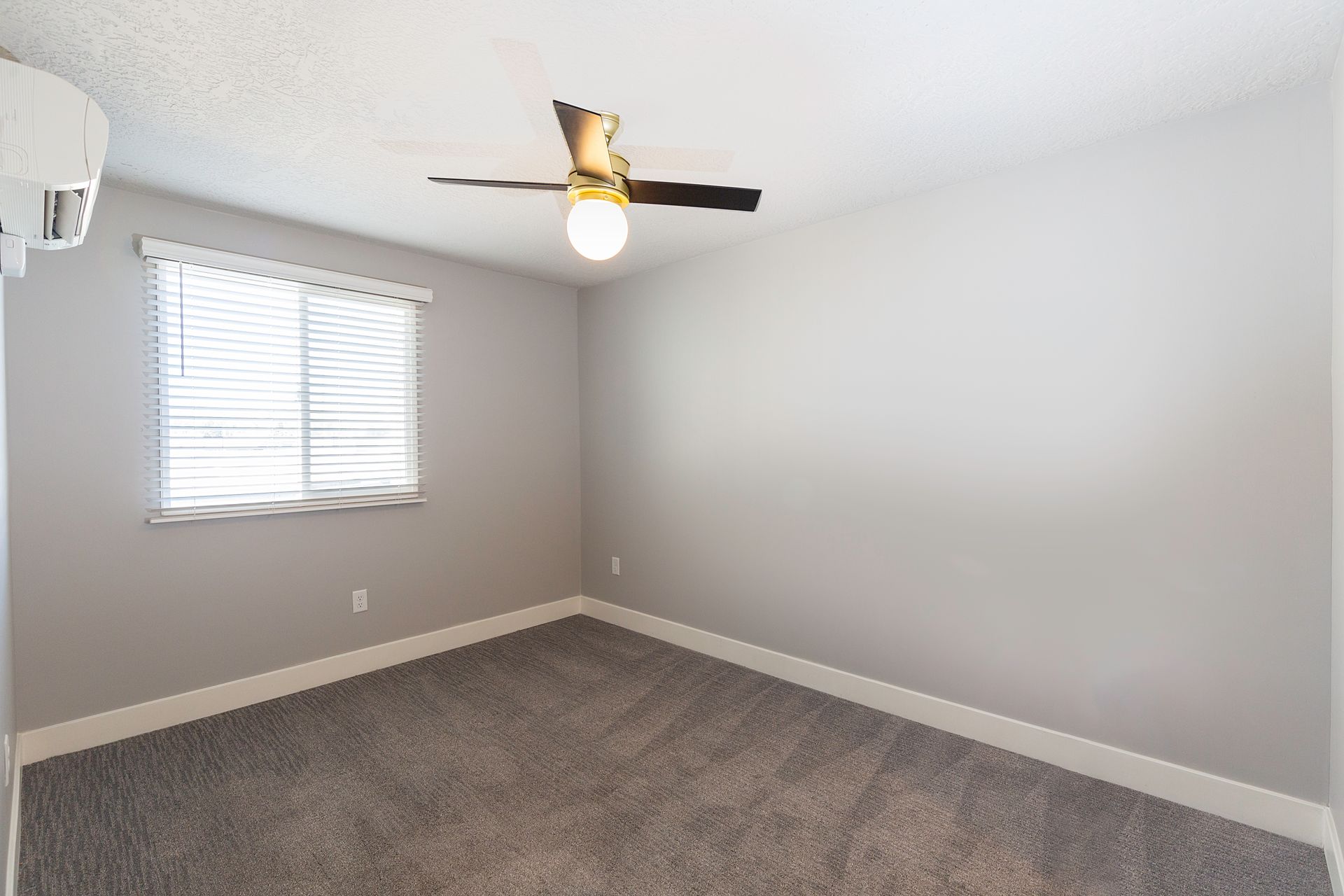 Empty bedroom with gray walls, window with blinds, ceiling fan, and gray carpet.