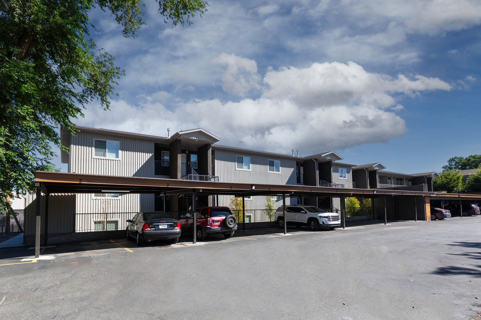 Apartment building with covered parking. Gray siding, tan carports, cars parked beneath. Sunny sky.