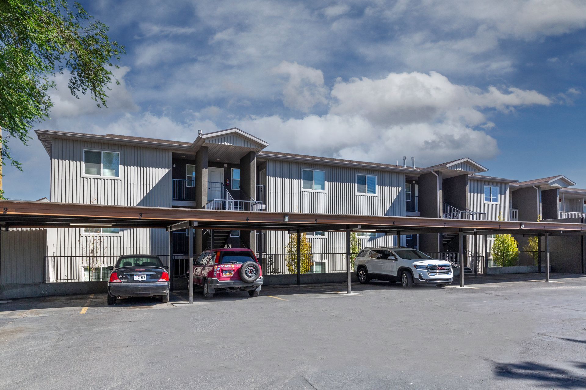 Apartment complex with covered parking; cars parked in front. Gray building, blue sky, clouds.