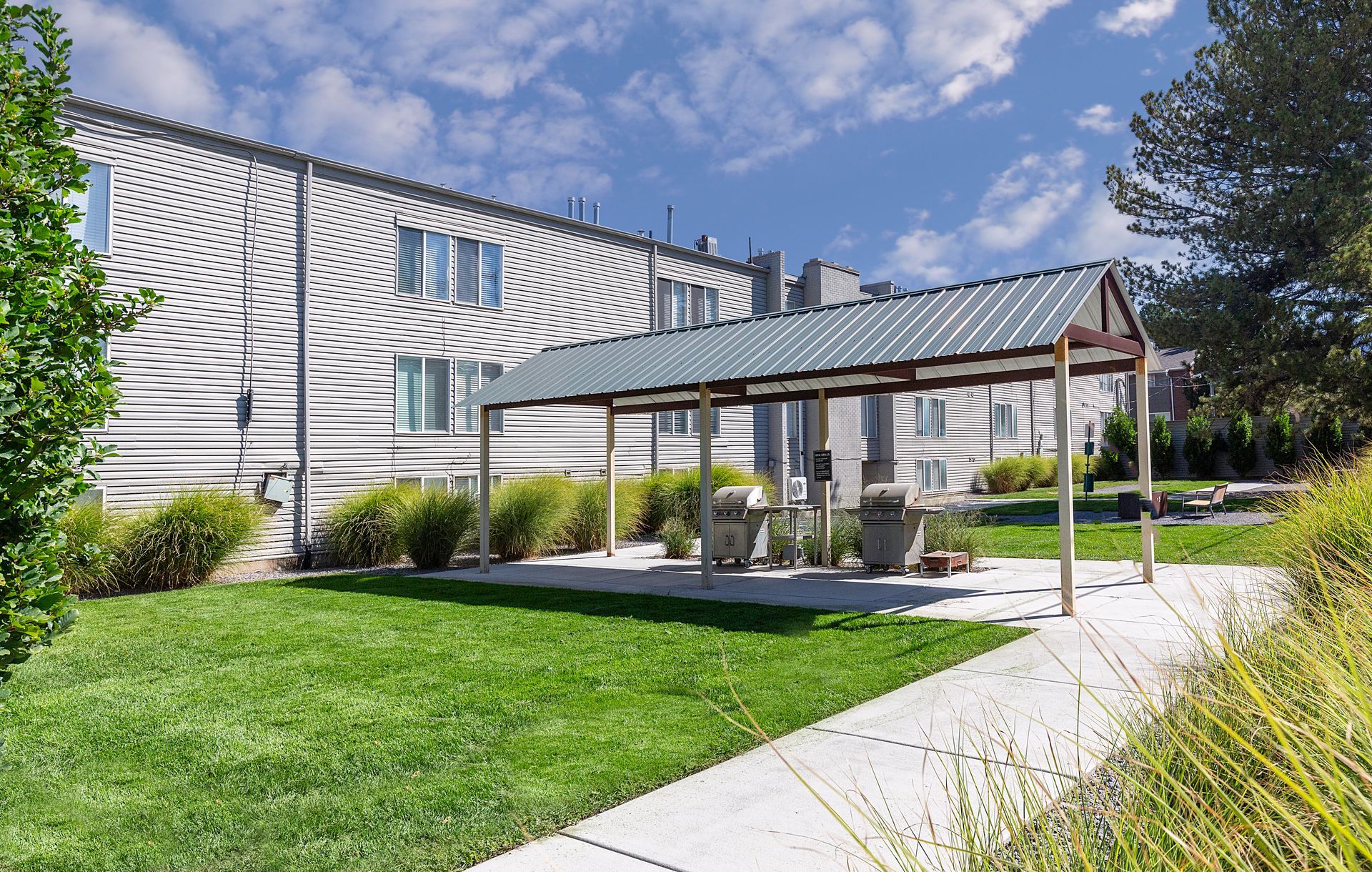 Grassy area with concrete path, covered grill area, and apartment building in the background on a sunny day.