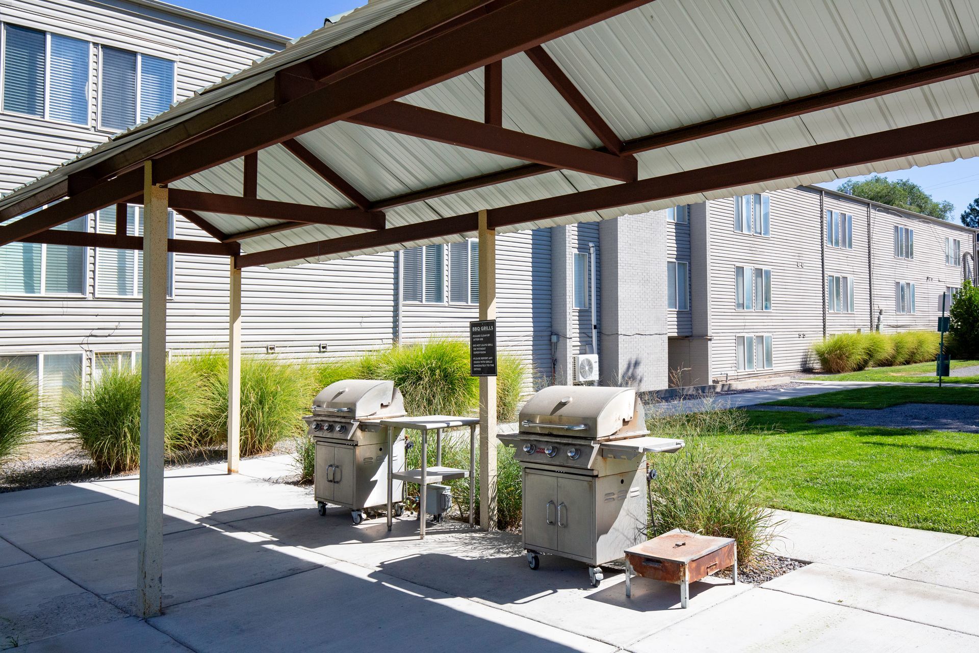 Covered outdoor grilling area with two grills, a small table, and apartment buildings in the background.