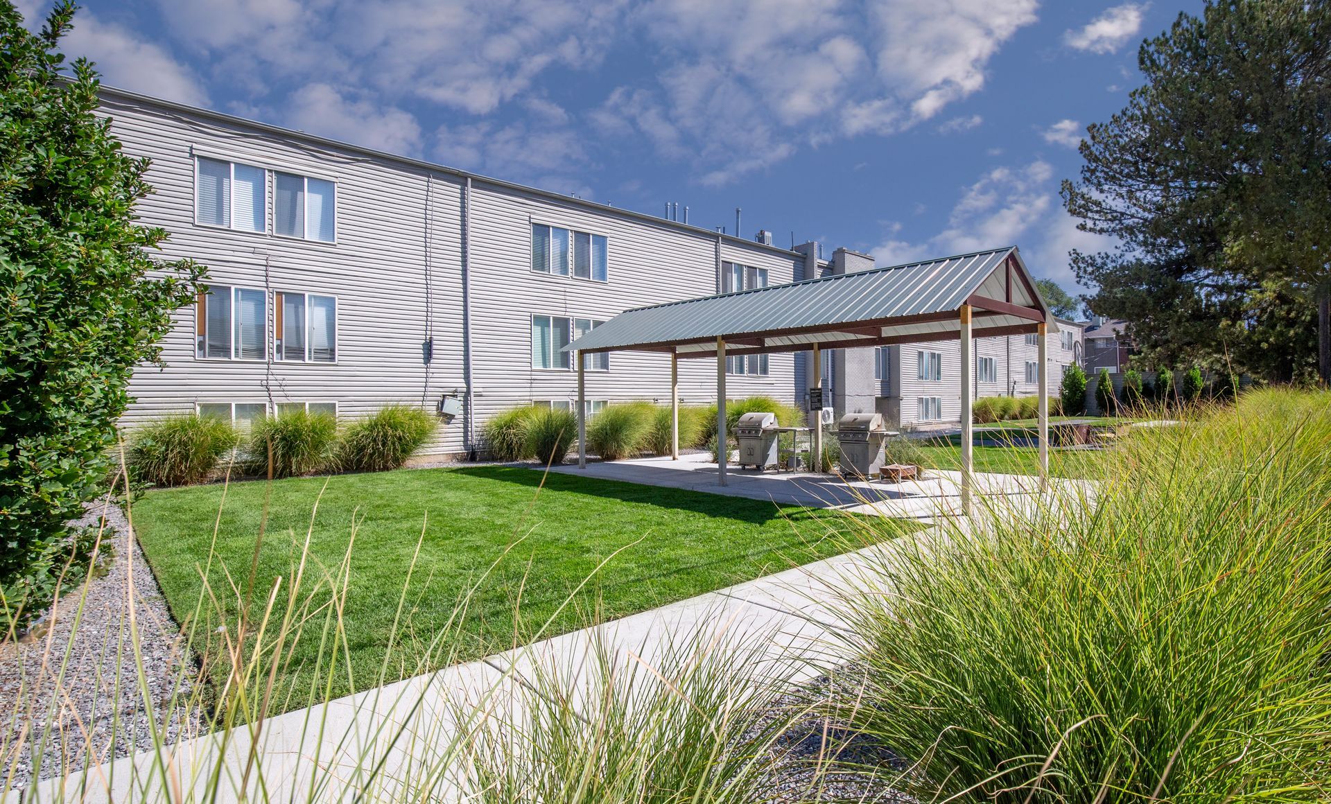 Grassy outdoor area with a covered seating area in front of a light-colored building under a partly cloudy sky.