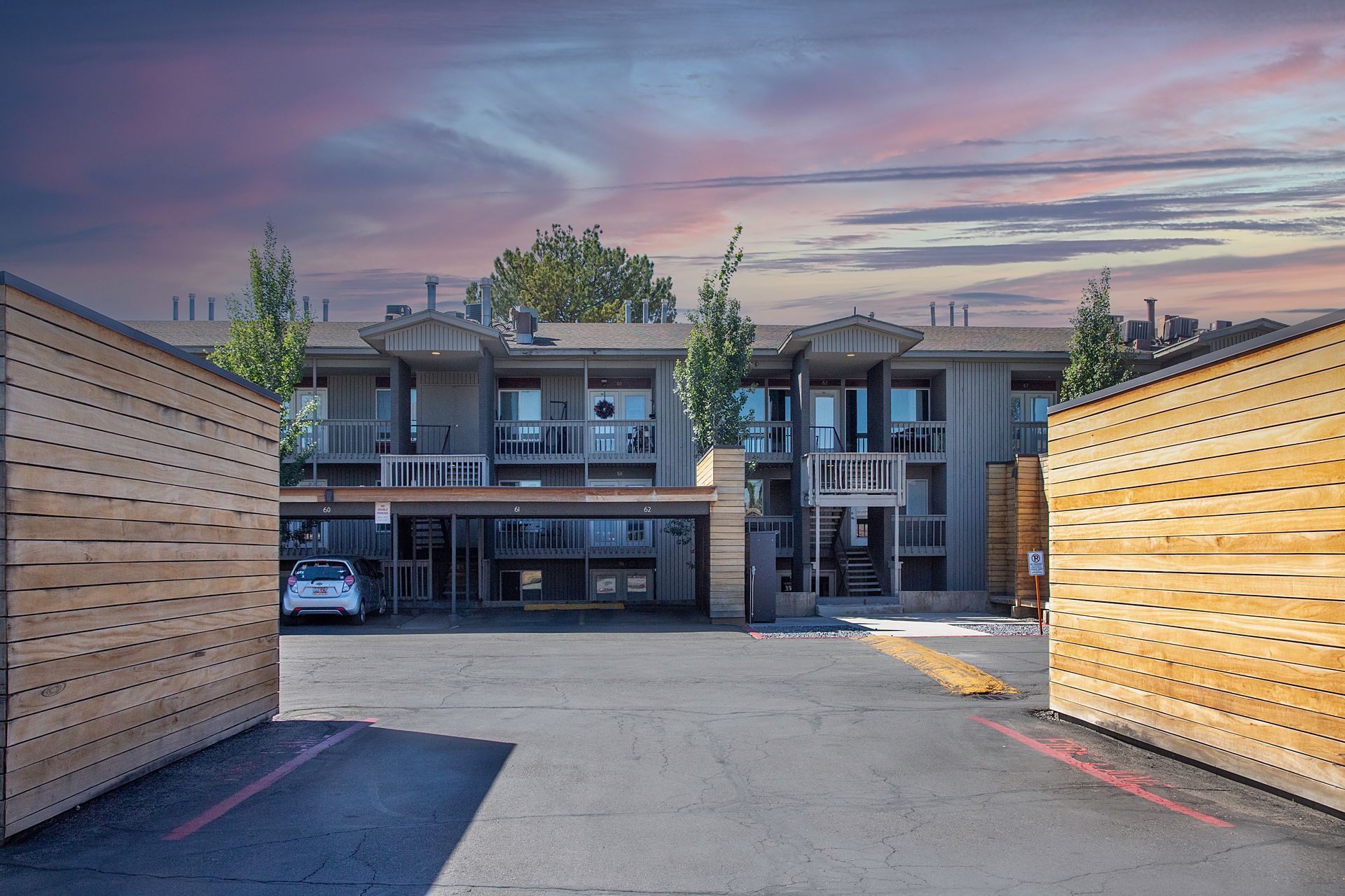 Apartment building with open wooden gates; car parked under a carport, evening sky.