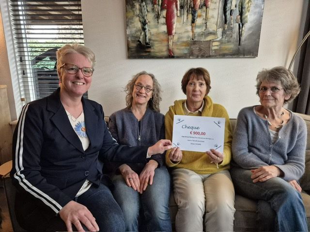 Vier vrouwen, met een grote cheque in hun handen, zitten lachend in een woonkamer.