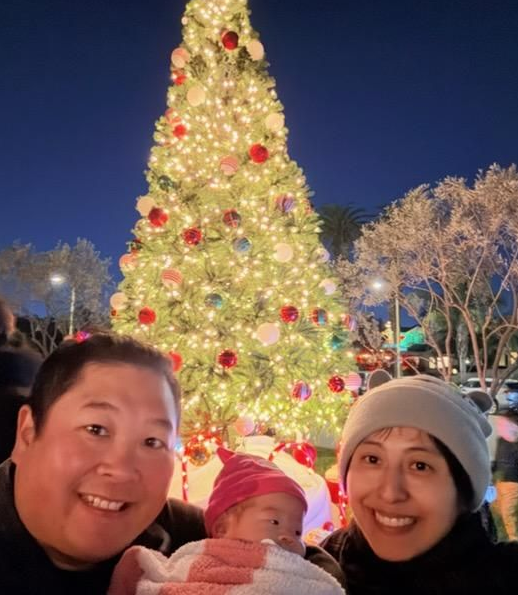 A couple smiles next to a baby in a pink hat, standing in front of a large, brightly lit Christmas tree at night.