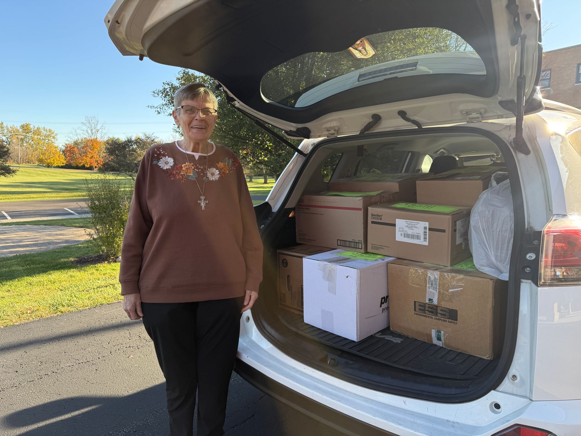 An individual stands beside an open car trunk filled with several cardboard boxes on a sunny day outdoors.