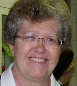 A person with curly hair and glasses smiling at the camera, wearing a white collared shirt.