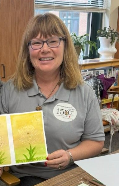 A person wearing glasses and a gray polo shirt holds up a small, yellow and green watercolor painting of a flower.