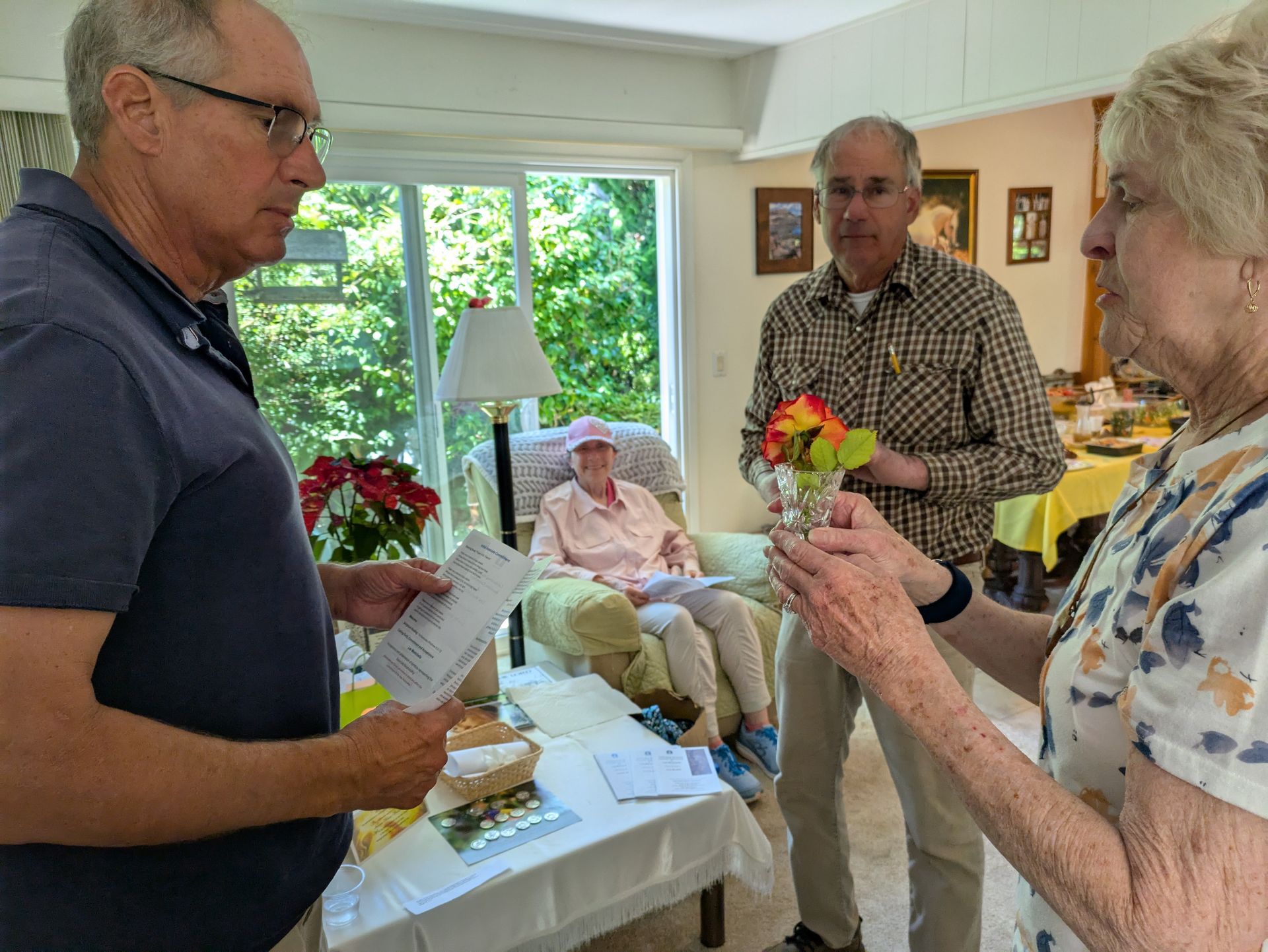Three people in a home; one reads a document, another holds a small floral arrangement, and one rests in an armchair.