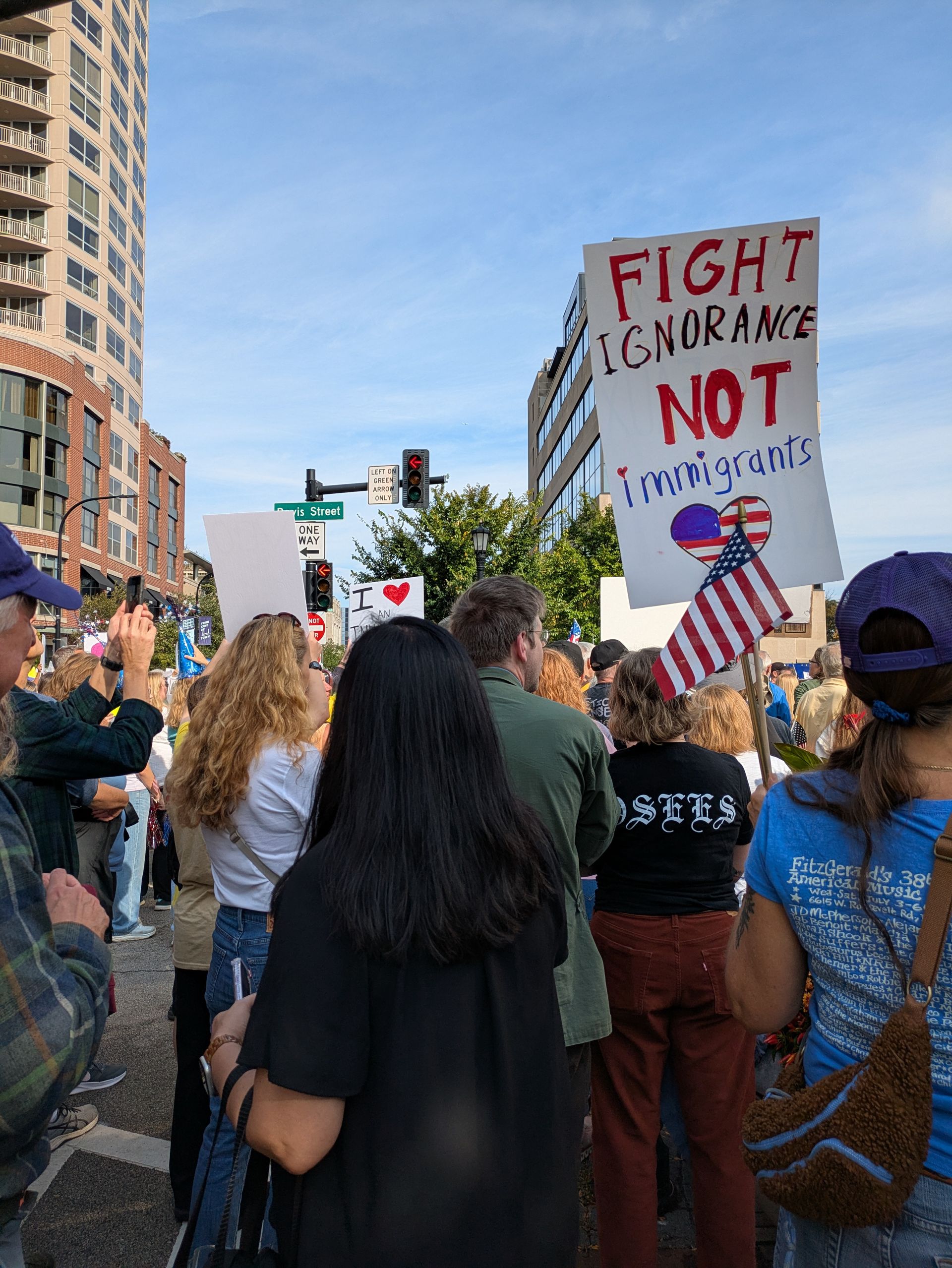 A crowd of people stands at a city intersection, one person holding a sign that says, 