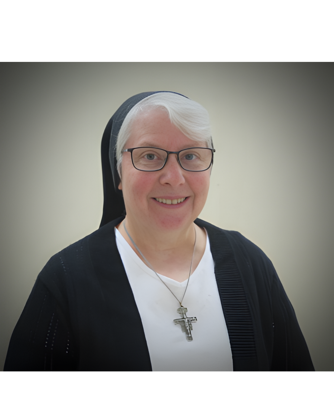 A smiling person wearing a black habit and veil, white undershirt, glasses, and a crucifix necklace against a gray background.
