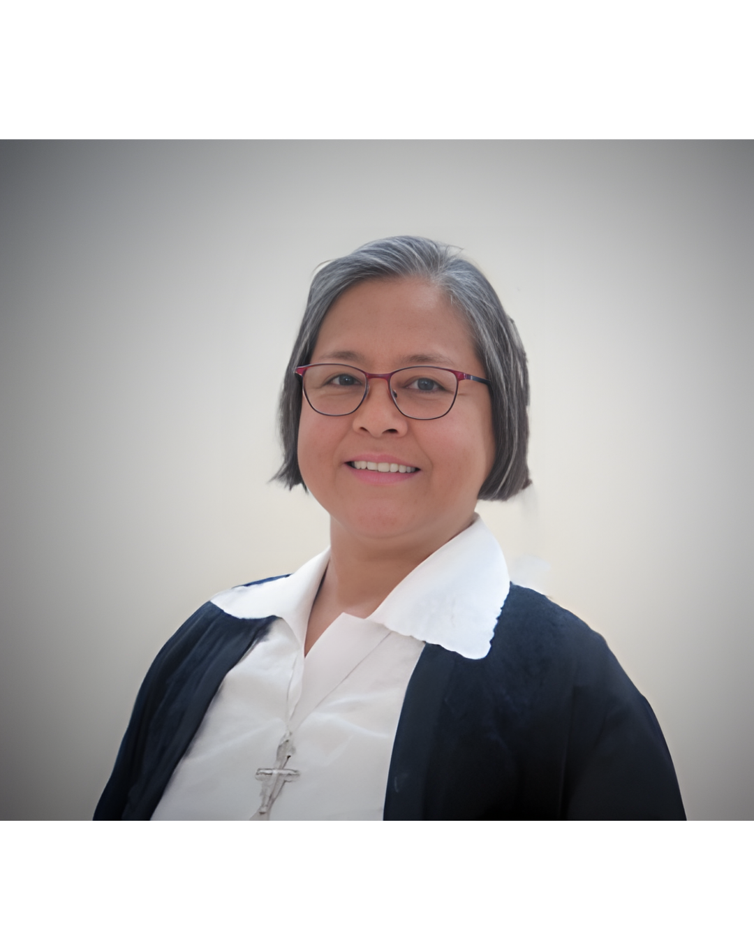 A person wearing glasses, a white collared shirt, a black cardigan, and a crucifix necklace, smiling against a plain wall.