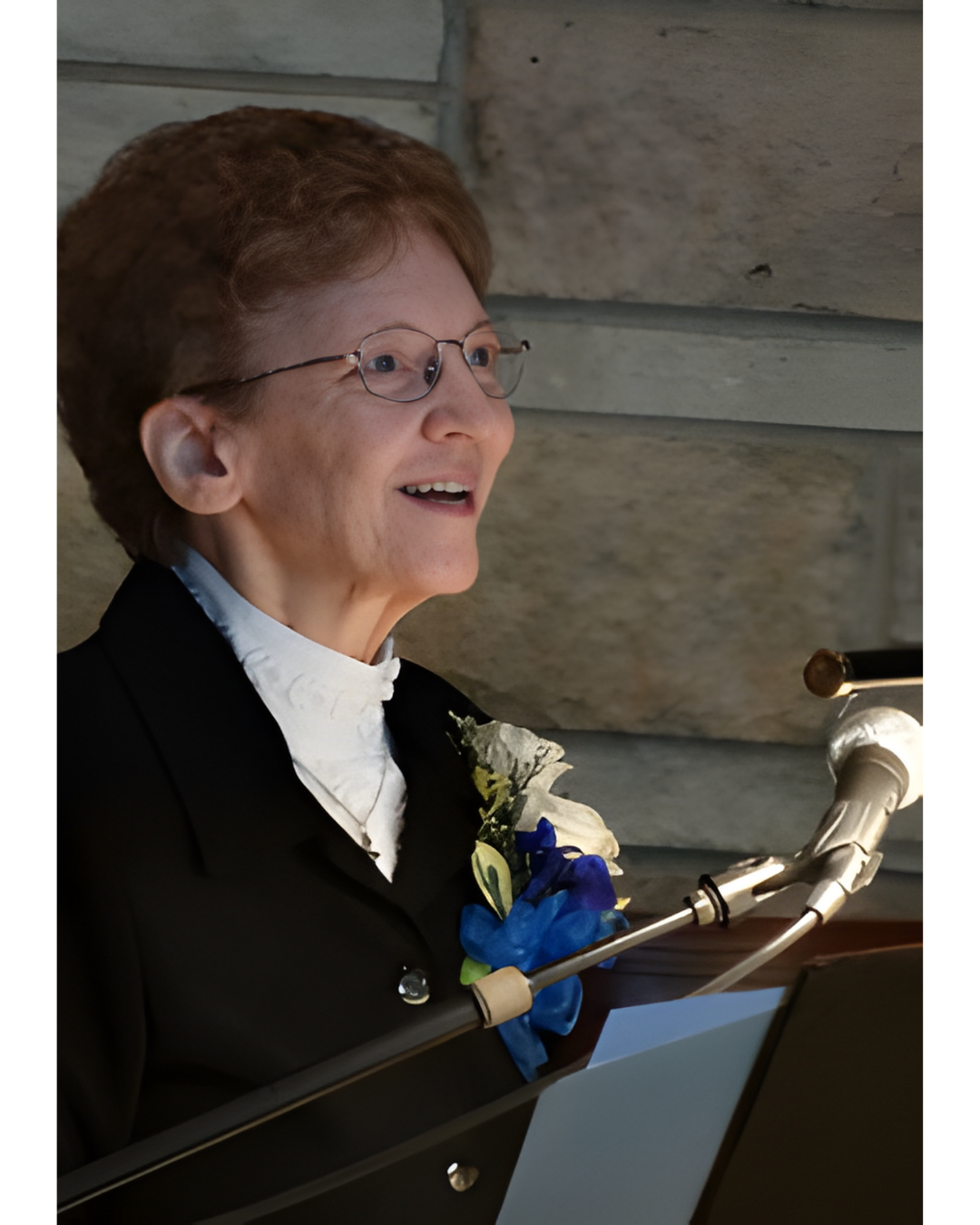 A person wearing glasses and a dark blazer speaks into a microphone in front of a stone wall.