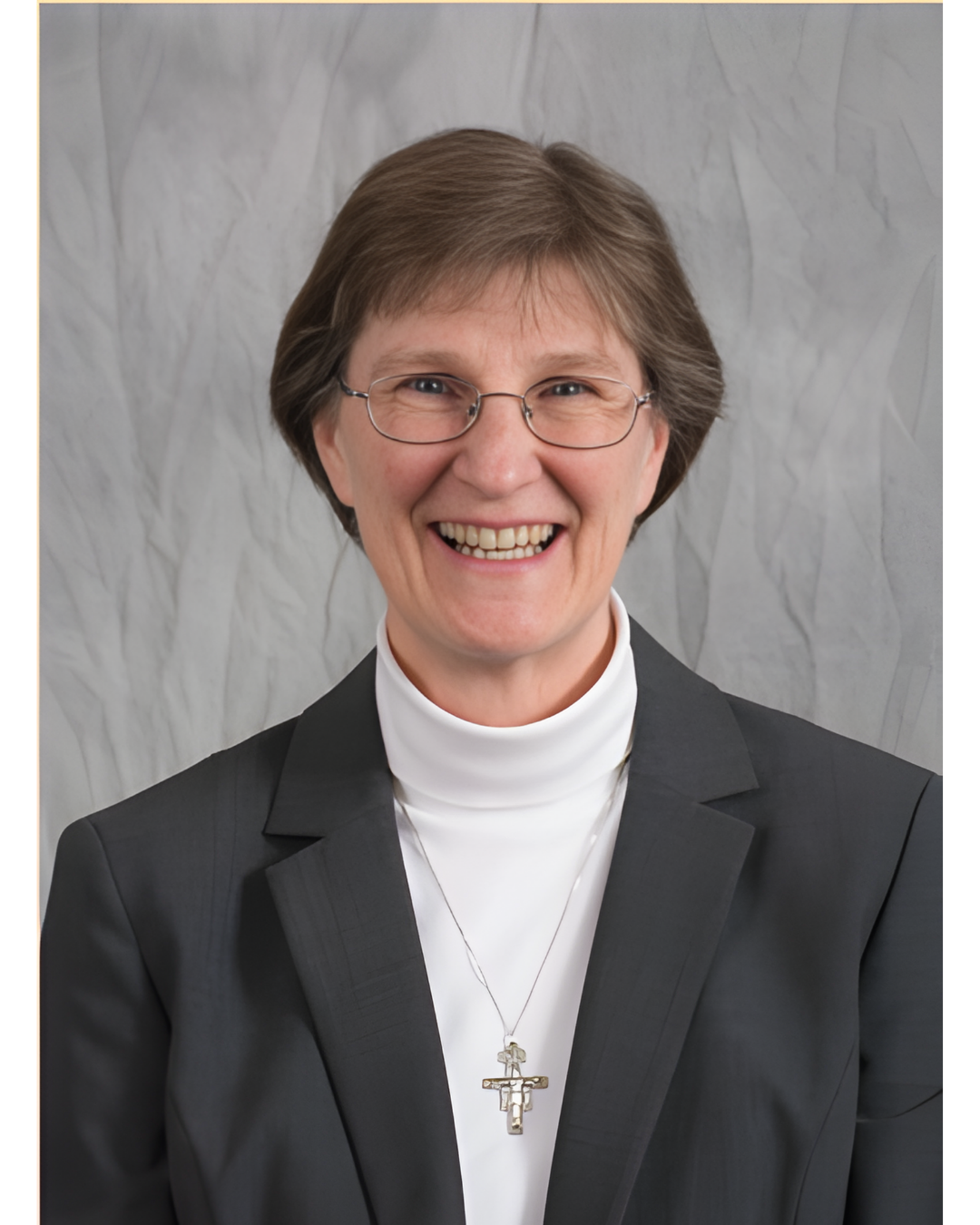 A smiling person with glasses, wearing a white turtleneck, a dark blazer, and a crucifix necklace against a gray backdrop.