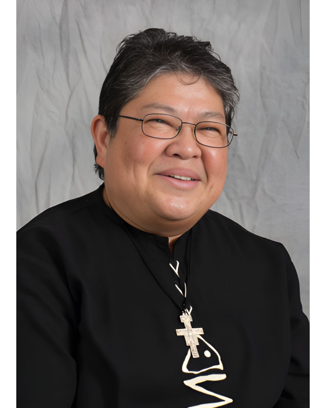 A person wearing glasses and a black shirt with a cross necklace smiles against a gray, textured background.