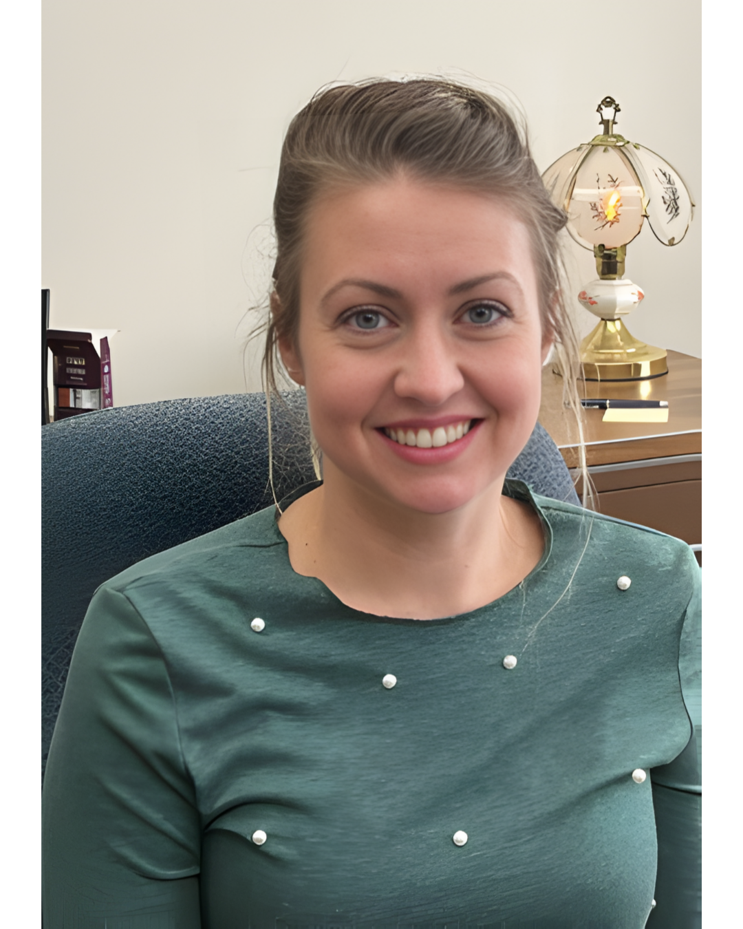 A smiling person wearing a green shirt with pearl-like accents, seated in an office with a lamp on a desk behind them.