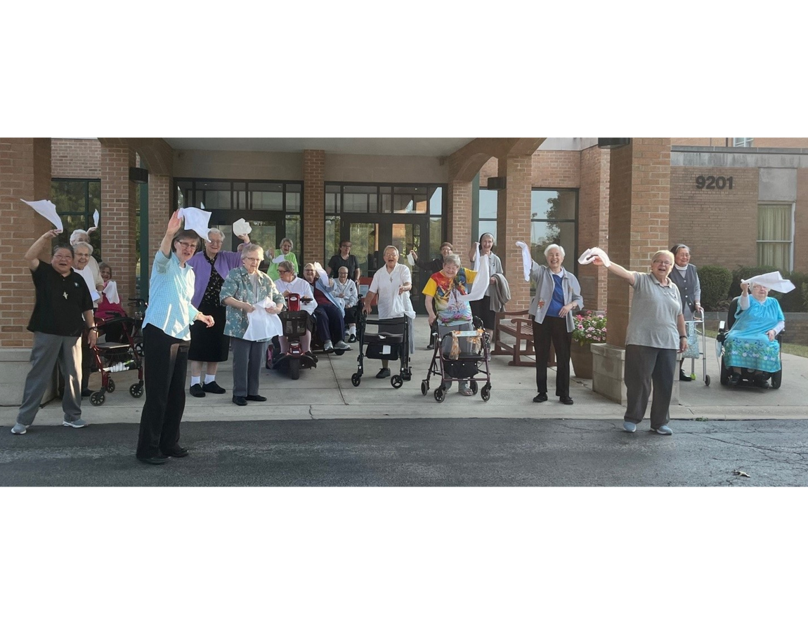 A group of people standing outside a brick building, waving white cloths and smiling.