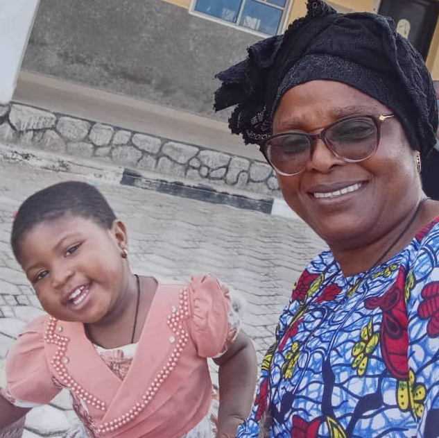 A smiling child and an adult in a patterned top and head covering pose together for a selfie outdoors.