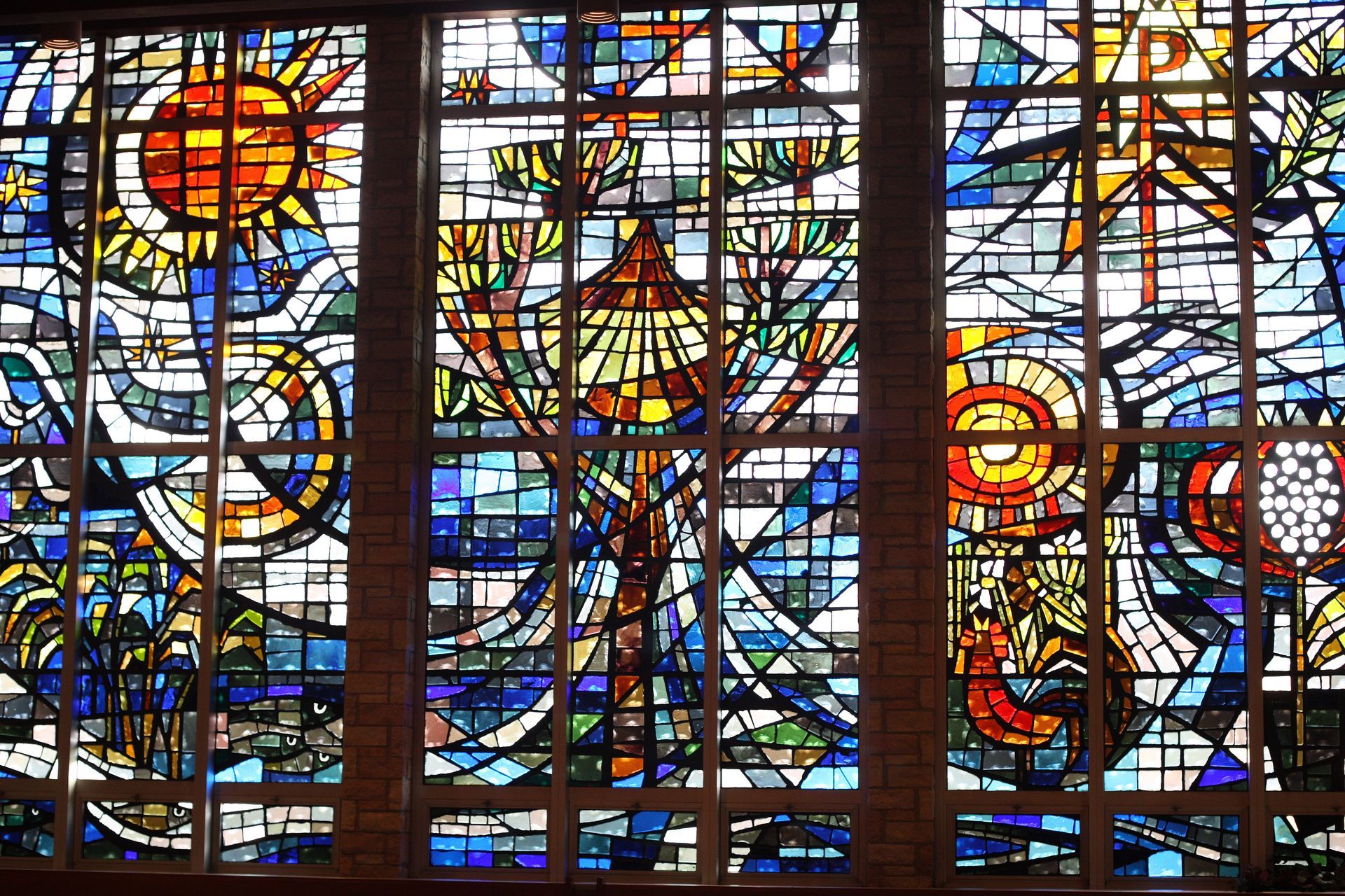 A vibrant, multi-paneled stained glass window featuring geometric shapes, a central tree, a bright sun, and a star.