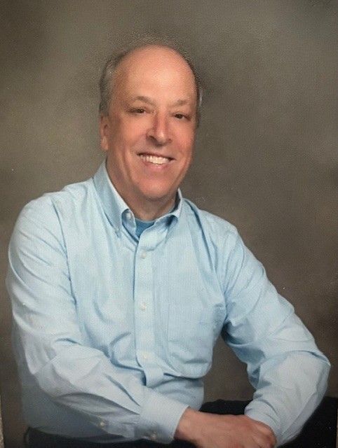 A person smiling while wearing a light blue, long-sleeved collared shirt against a gray backdrop.