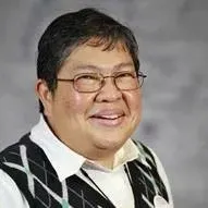 A smiling person wearing glasses and a dark, patterned V-neck sweater over a white collared shirt against a gray background.