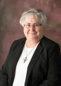 A person wearing glasses, a dark blazer, and a cross necklace smiles against a mottled, dark-red background.